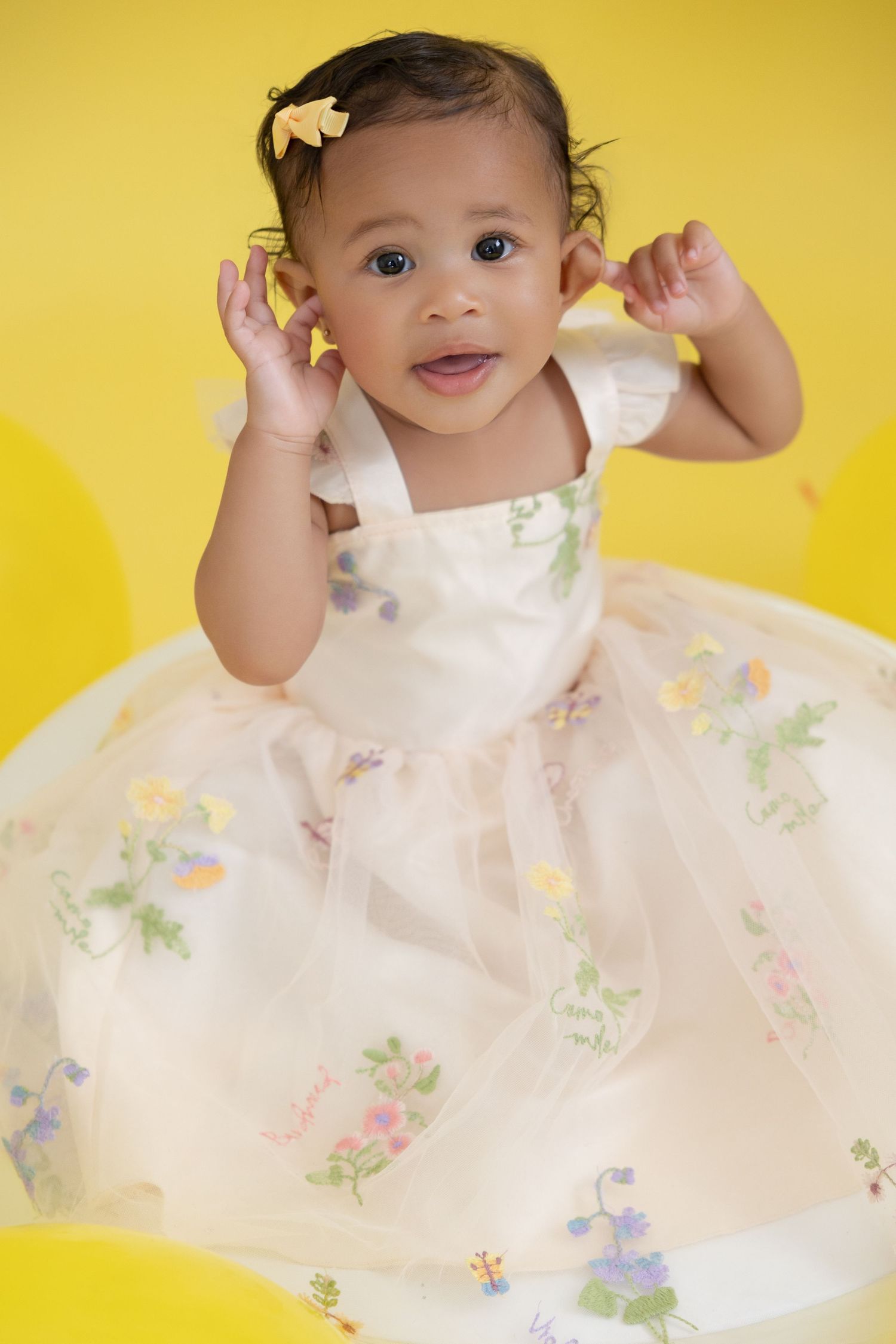Adorable baby girl in a floral white dress with a yellow bow sits against a bright yellow background.