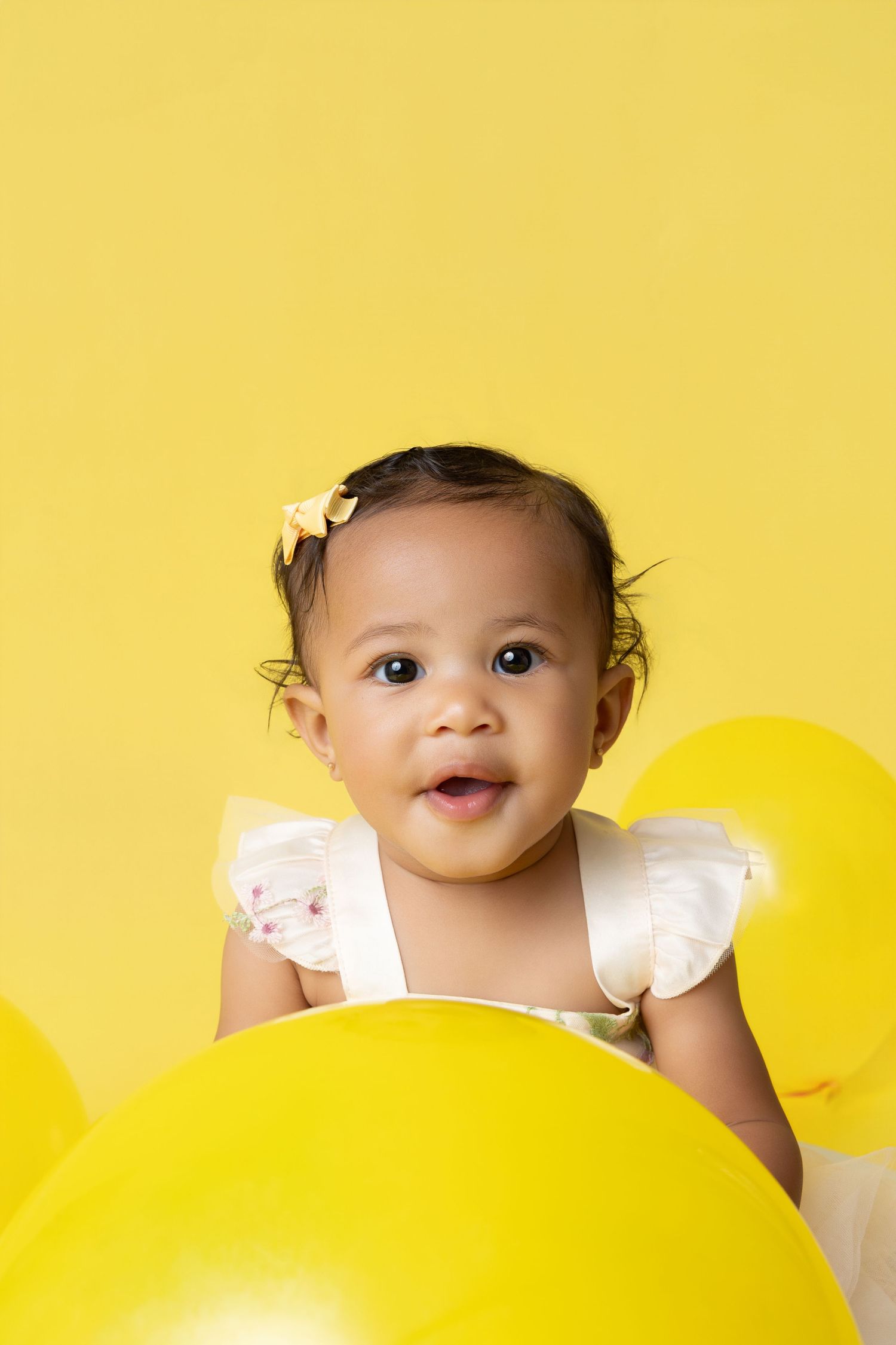Adorable baby girl in white dress surrounded by yellow balloons against a bright yellow background.