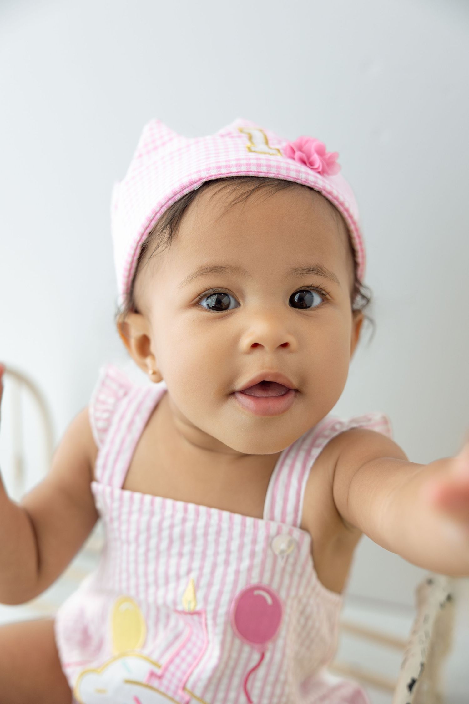 Adorable baby girl in pink striped romper and matching cat ear hat with floral detail reaching out toward camera.