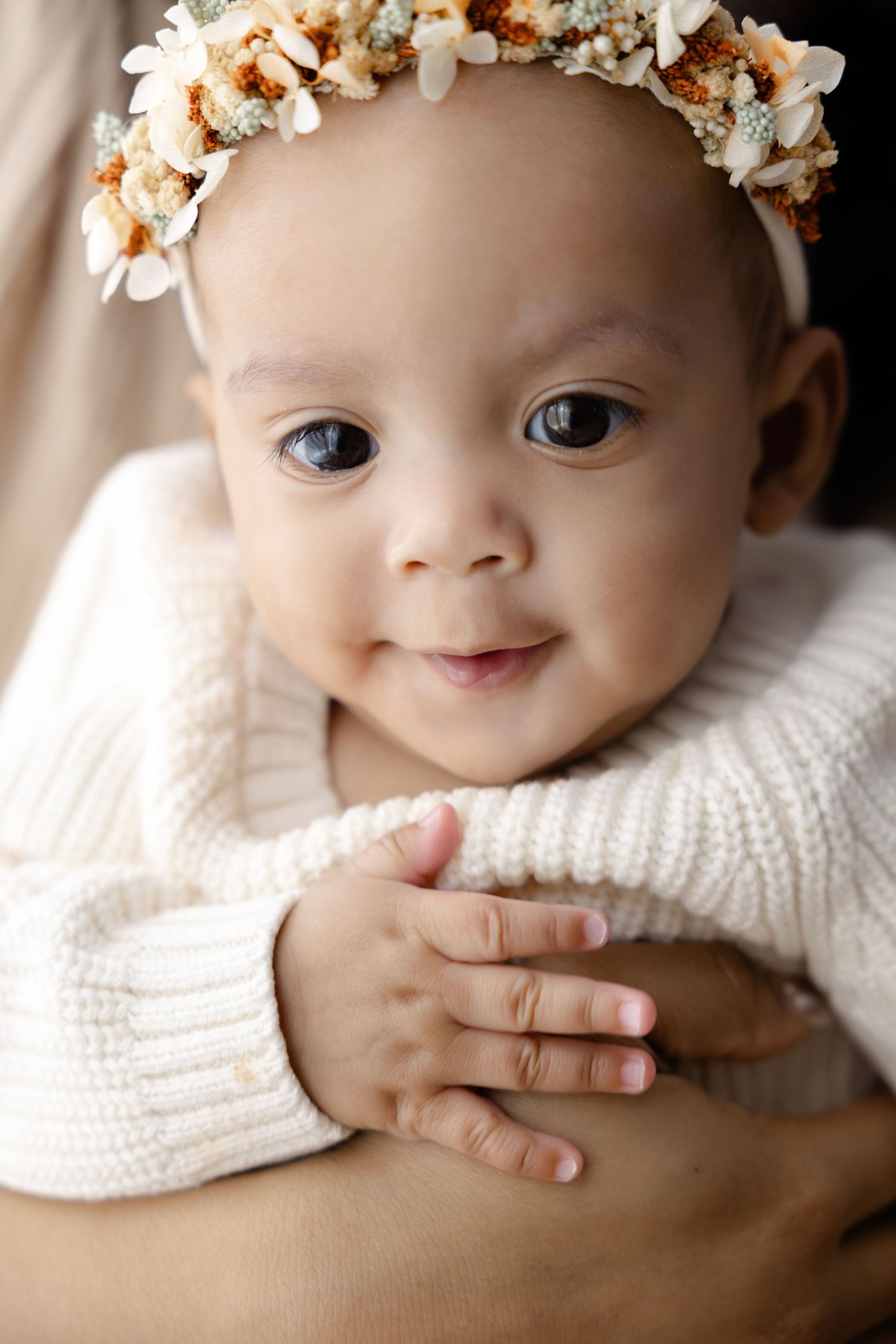 A baby wearing a white knit sweater and delicate floral headband smiles sweetly while lying down.