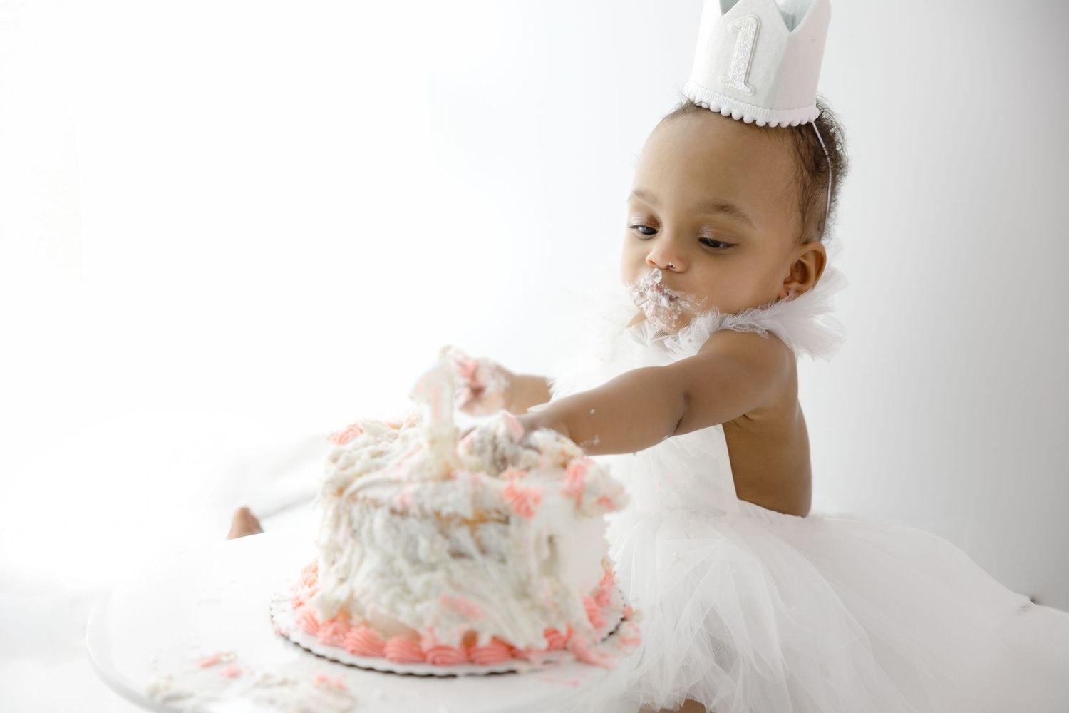 A small figure in white party attire and paper crown digs into a white and pink birthday cake, making a delightful mess.