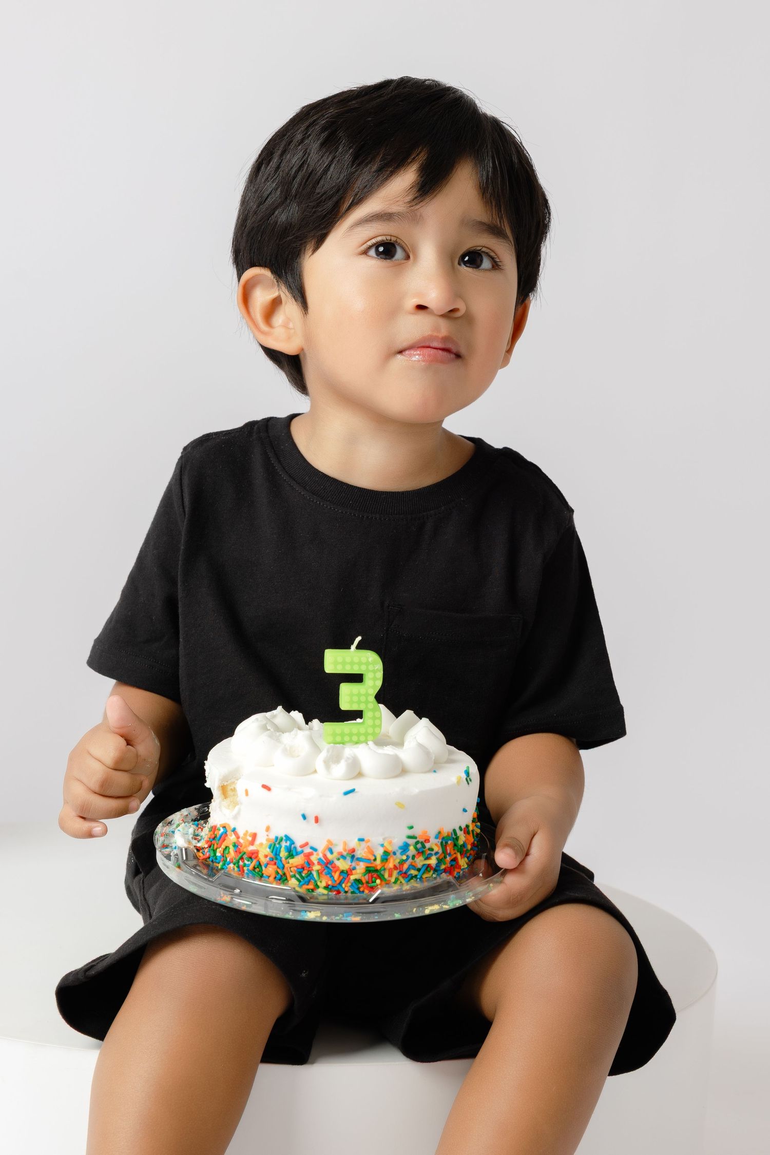 Young child in black shirt holding white frosted birthday cake with colorful sprinkles against white background.