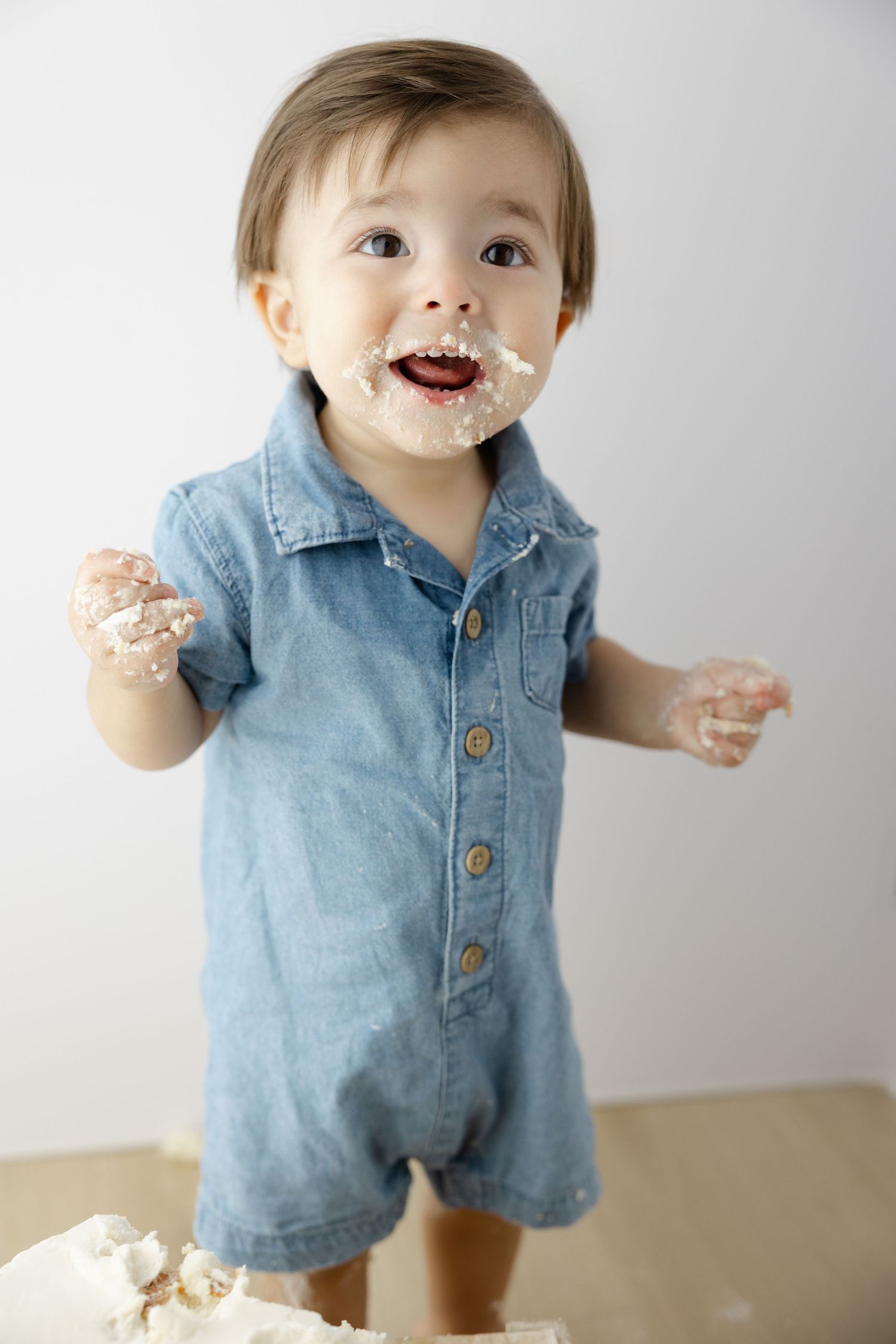 Baby wearing denim romper plays with cake frosting while making a joyful expression against white background.