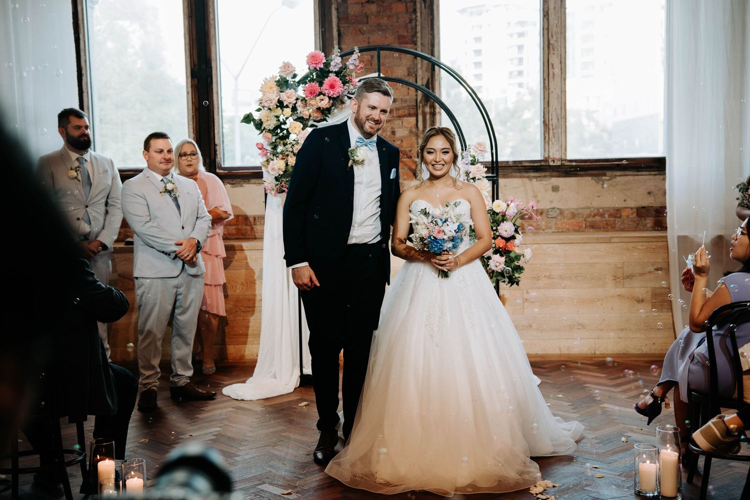 An elegant wedding ceremony in a rustic venue with brick walls and floral arch decor.