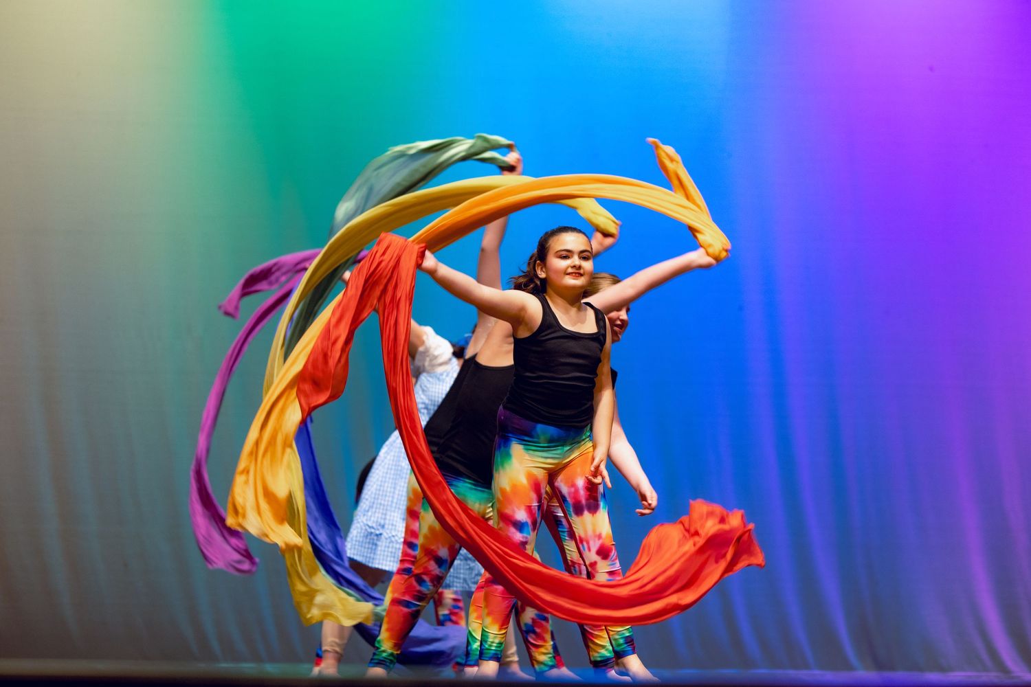 Dancer performs with colorful flowing ribbons against a rainbow gradient background.