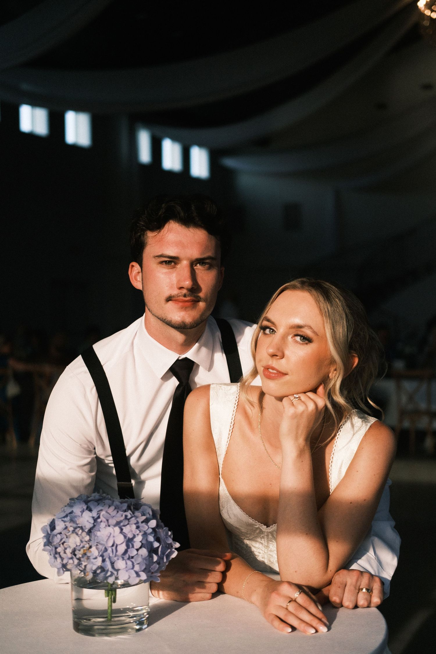 Newlywed couple sitting closely at a decorated dinner table, smiling and looking towards on wedding reception