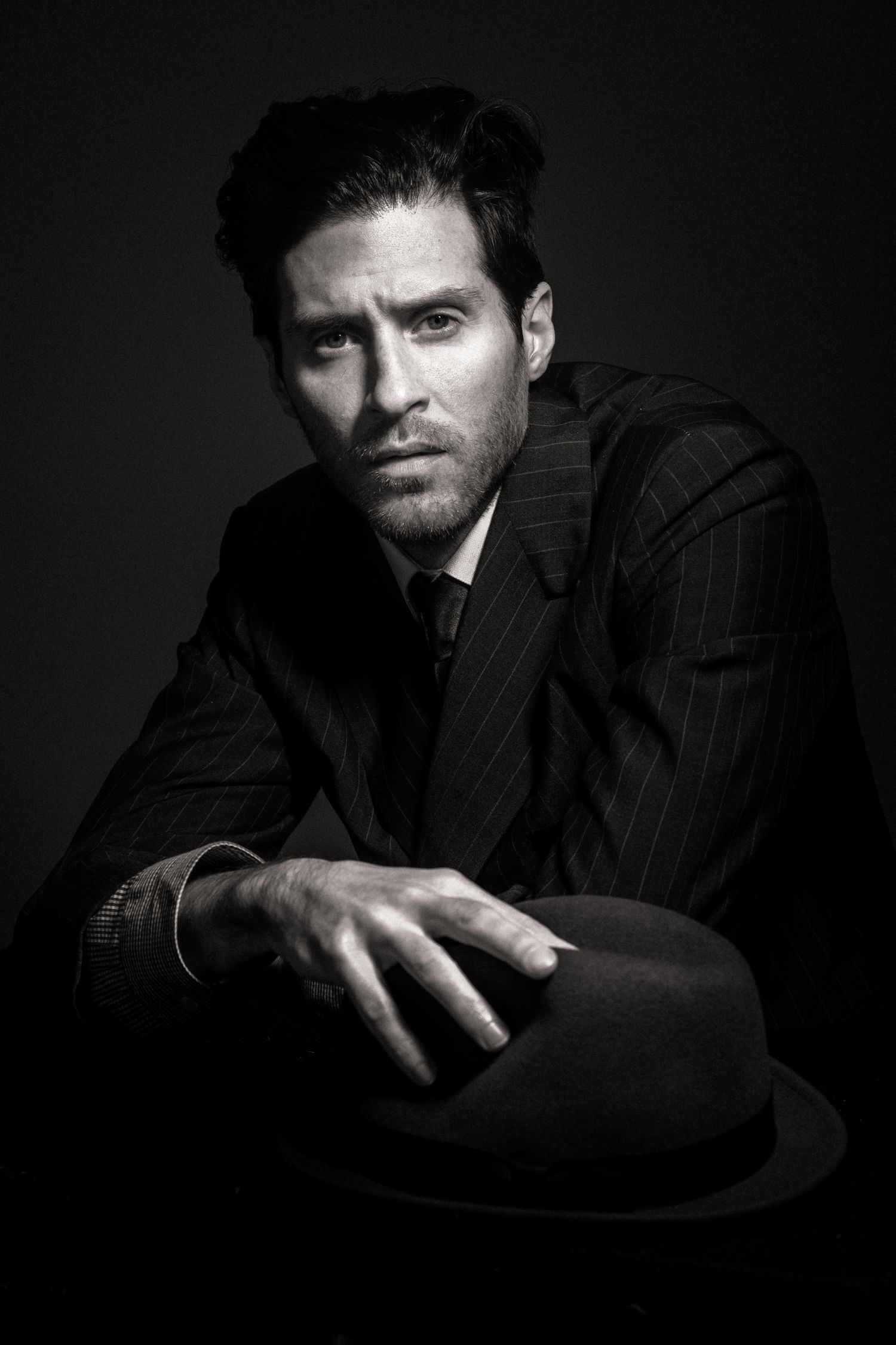 Dramatic black and white portrait of a man in pinstripe suit holding a fedora hat against dark background.