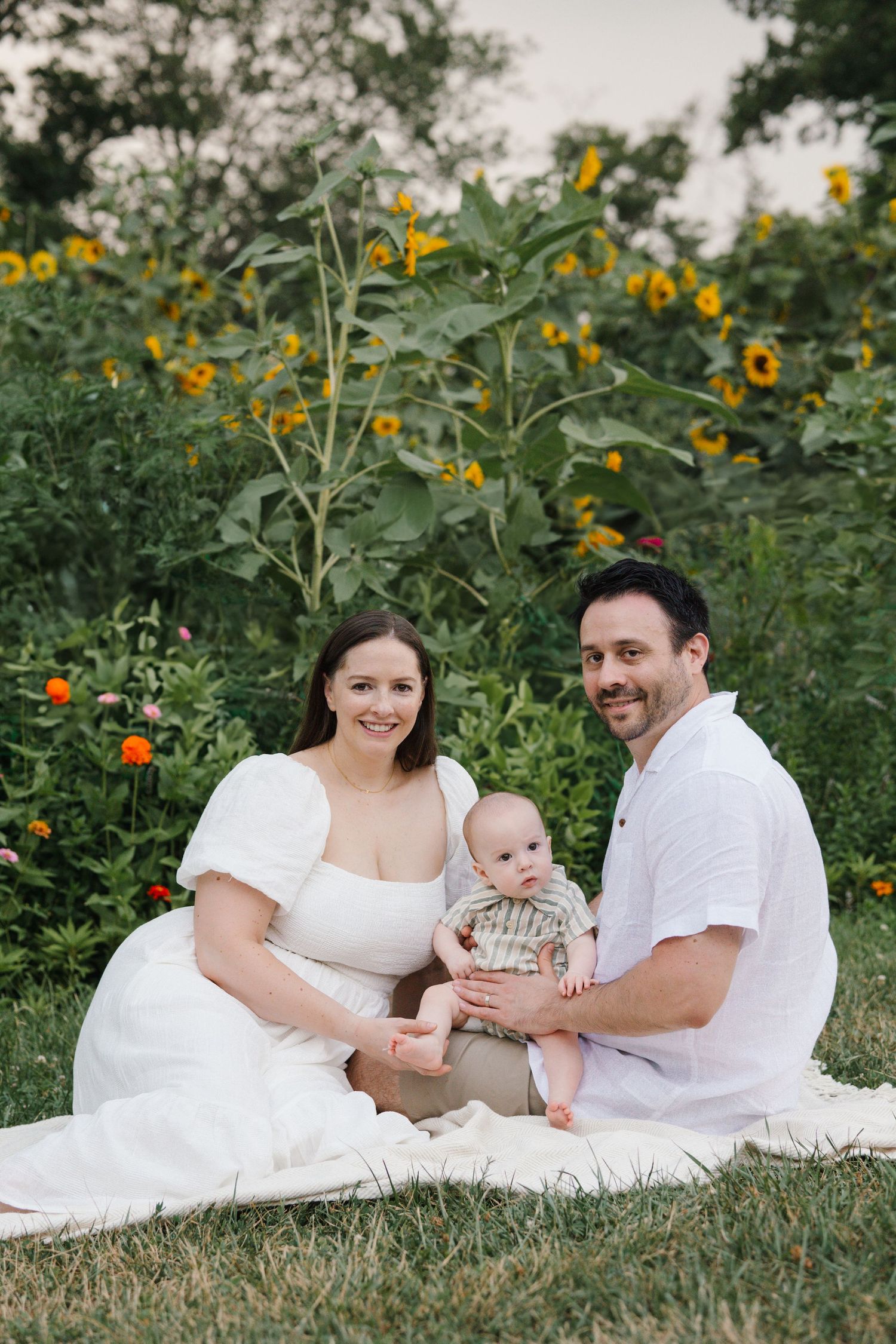A family sits together on a white blanket in front of yellow sunflowers blooming in the community gardens at Barclay Farmstead.