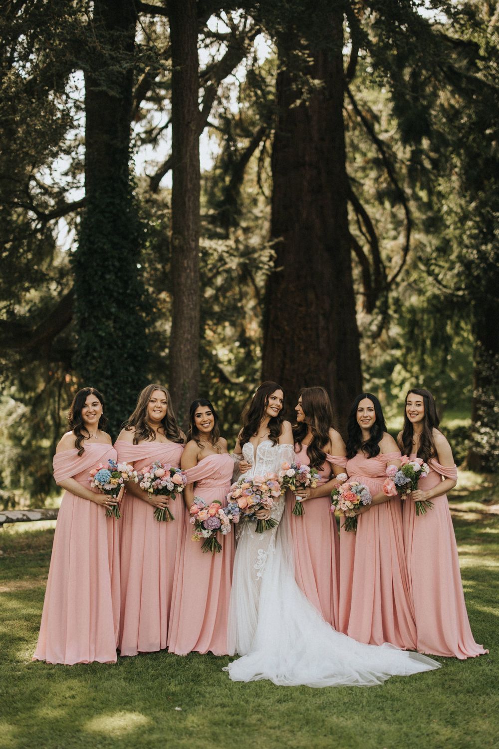 Wedding party group in pink bridesmaid dresses and white bridal gown posing on lawn with trees in background at Askham Hall.