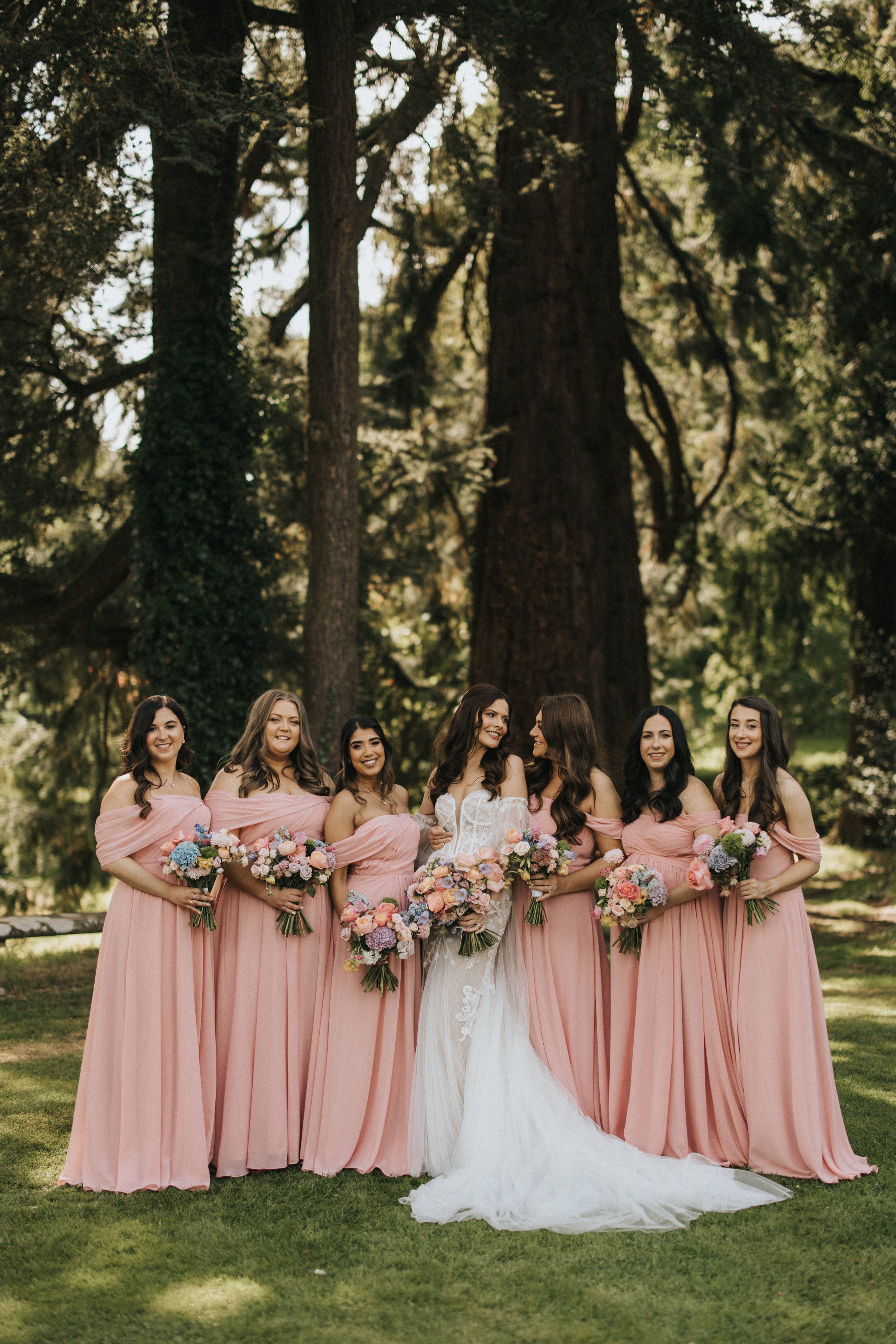 Wedding party group in pink bridesmaid dresses and white bridal gown posing on lawn with trees in background at Askham Hall.