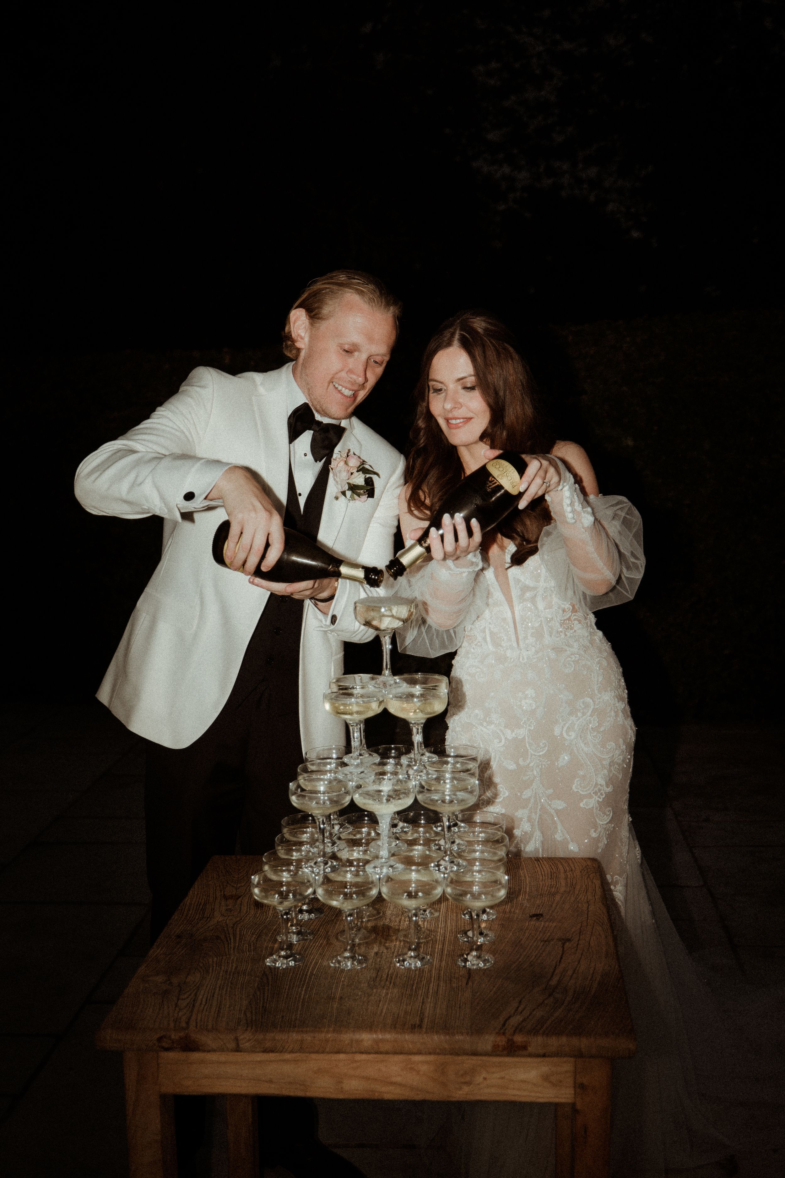 Couple pouring champagne into tower of vintage glasses on wooden table in dark setting at Askham Hall.