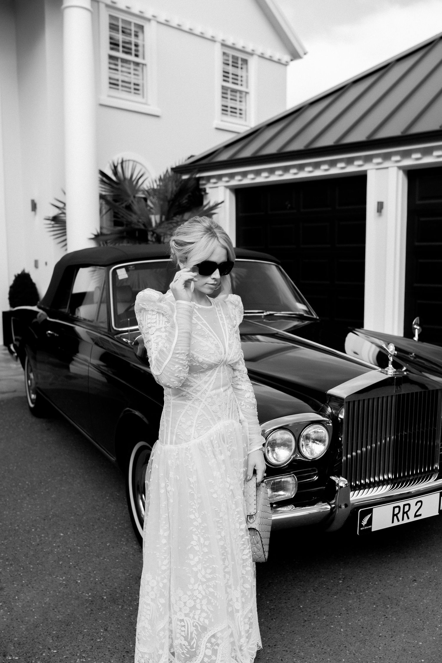 A bride in an elegant lace wedding dress stands beside a classic black Rolls Royce outside a white building.