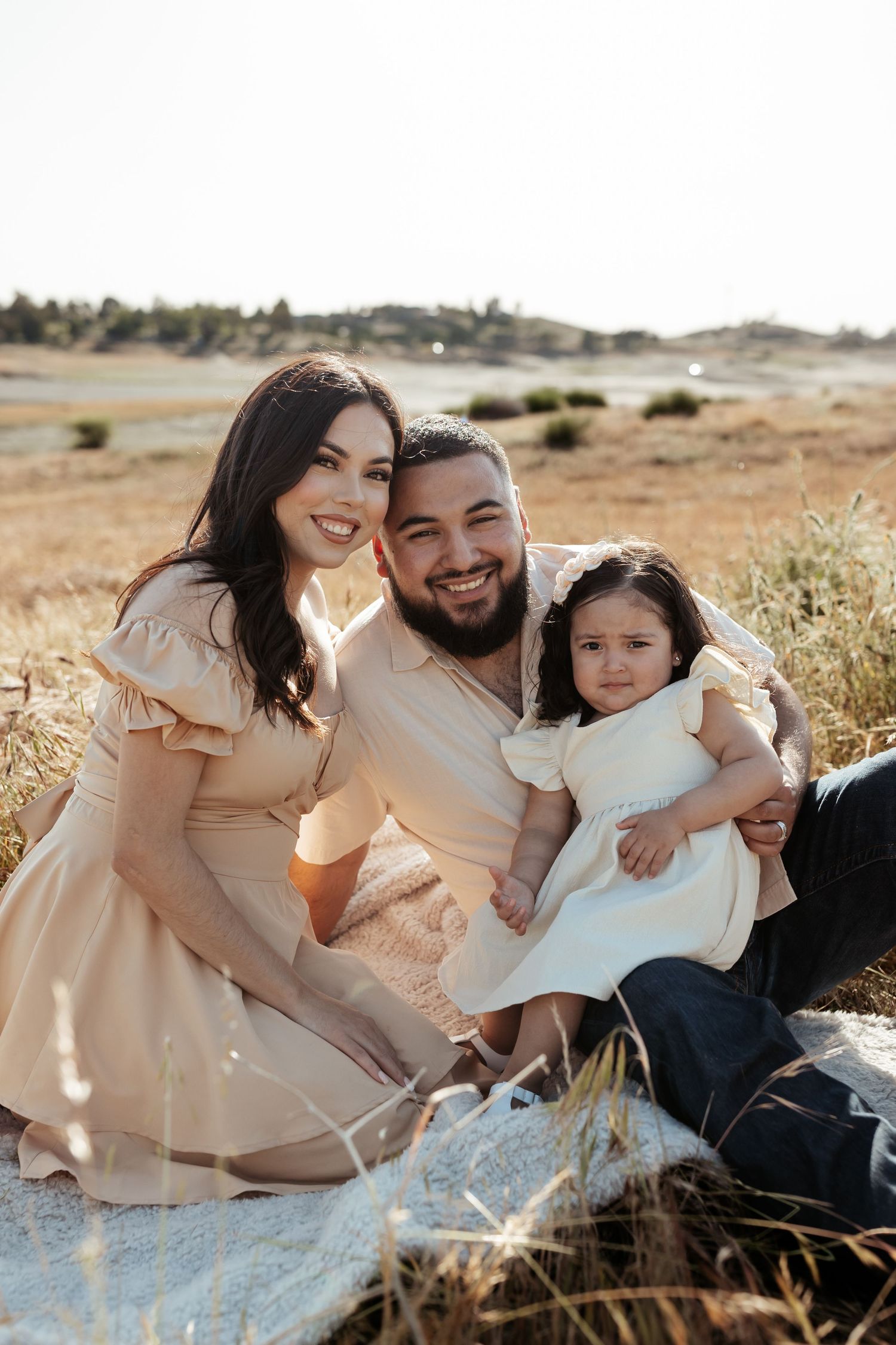 a couple and child sitting in a golden field of grass outdoors smiling at the camera