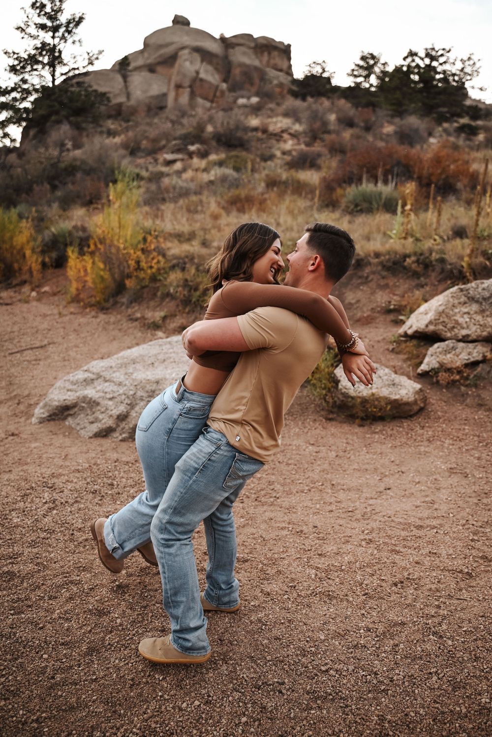 Playful couple embracing on dirt trail with rocky mountain backdrop.