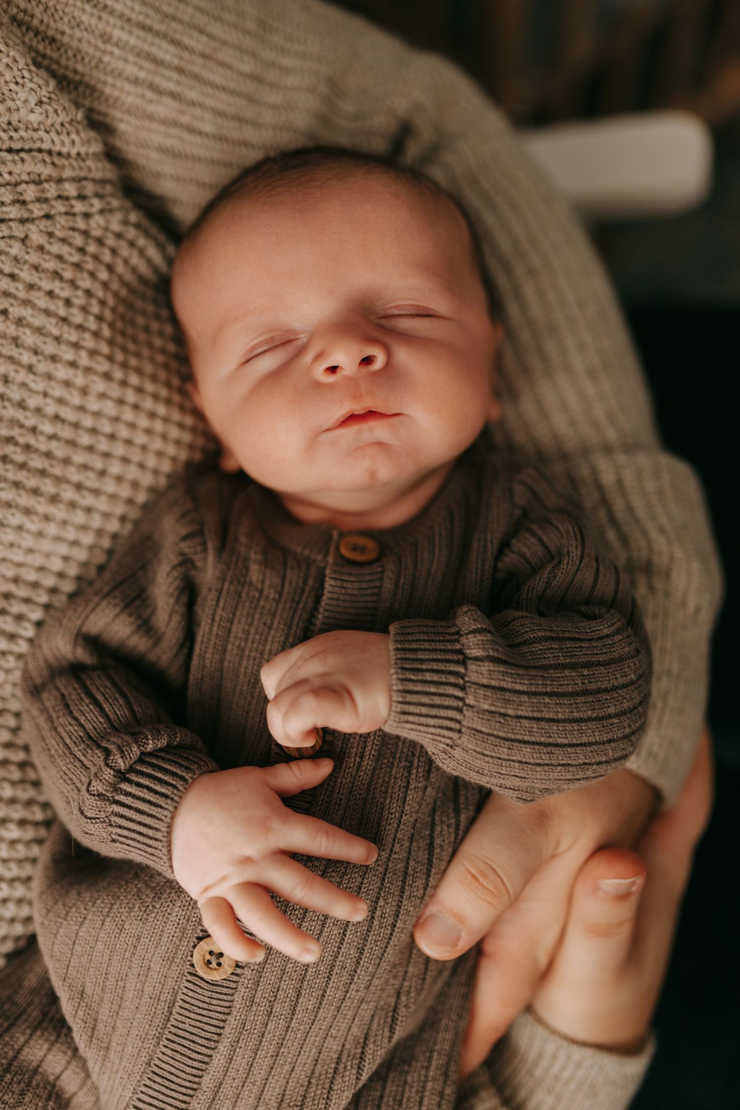 Sleeping newborn baby in knit olive cardigan cradled in parents arms in warm cozy setting.
