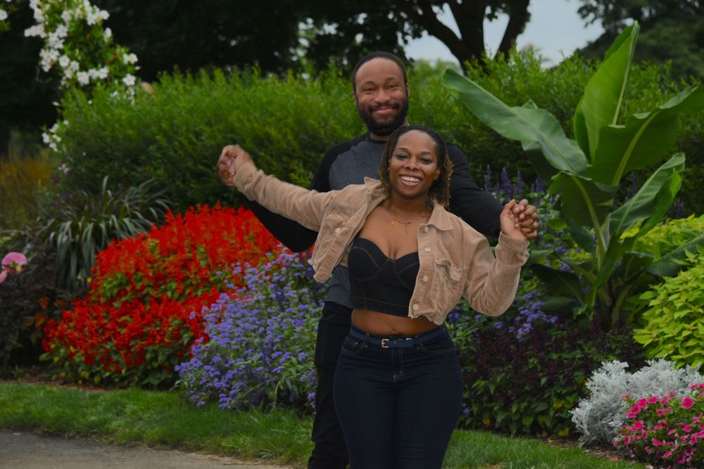 A joyful couple poses in a garden with vibrant red flowers and lush greenery.