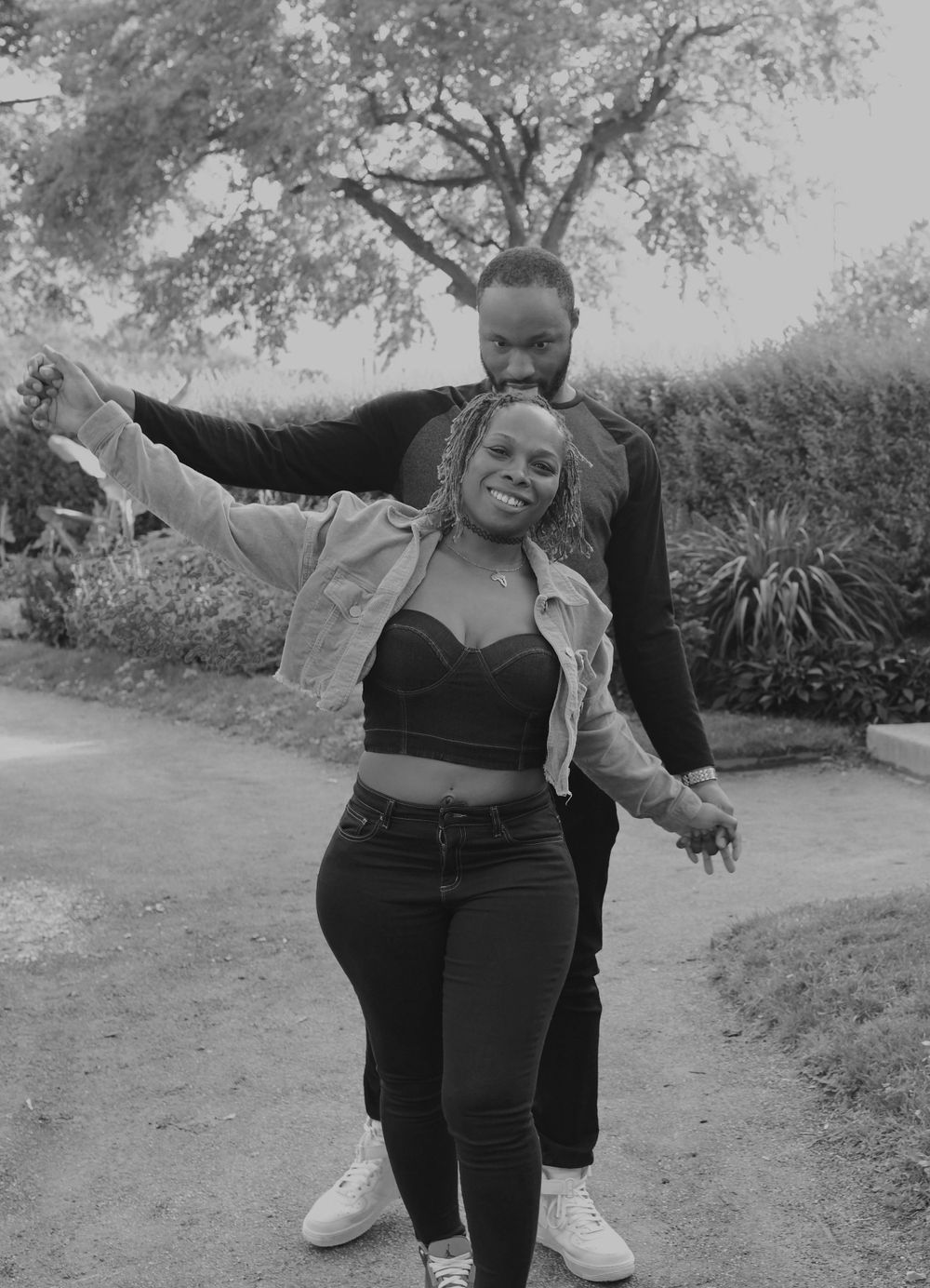 A couple posed outdoors in a playful dancing pose along a garden path in black and white.
