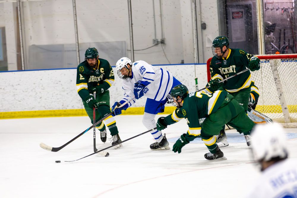 Hockey player in white jersey battles against multiple defenders in green uniforms.