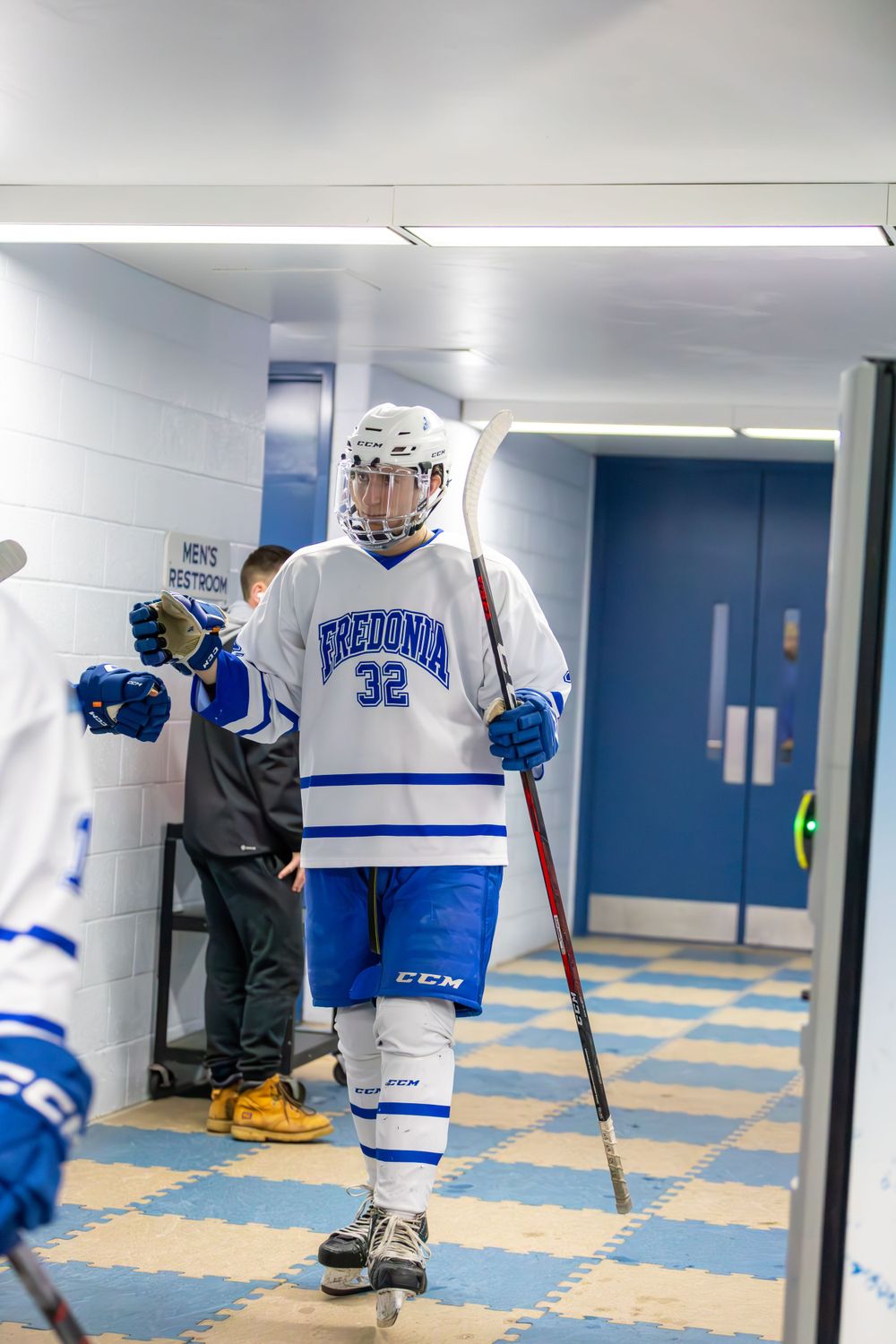 Hockey players in Fredonia uniforms walk through a locker room hallway with blue and yellow floor tiles.