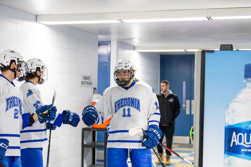 Hockey players in blue and white uniforms prepare in a well-lit facility hallway.