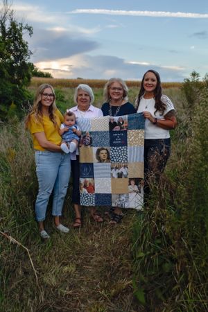 Four generations display a handmade patchwork quilt outdoors at sunset in a grassy field.
