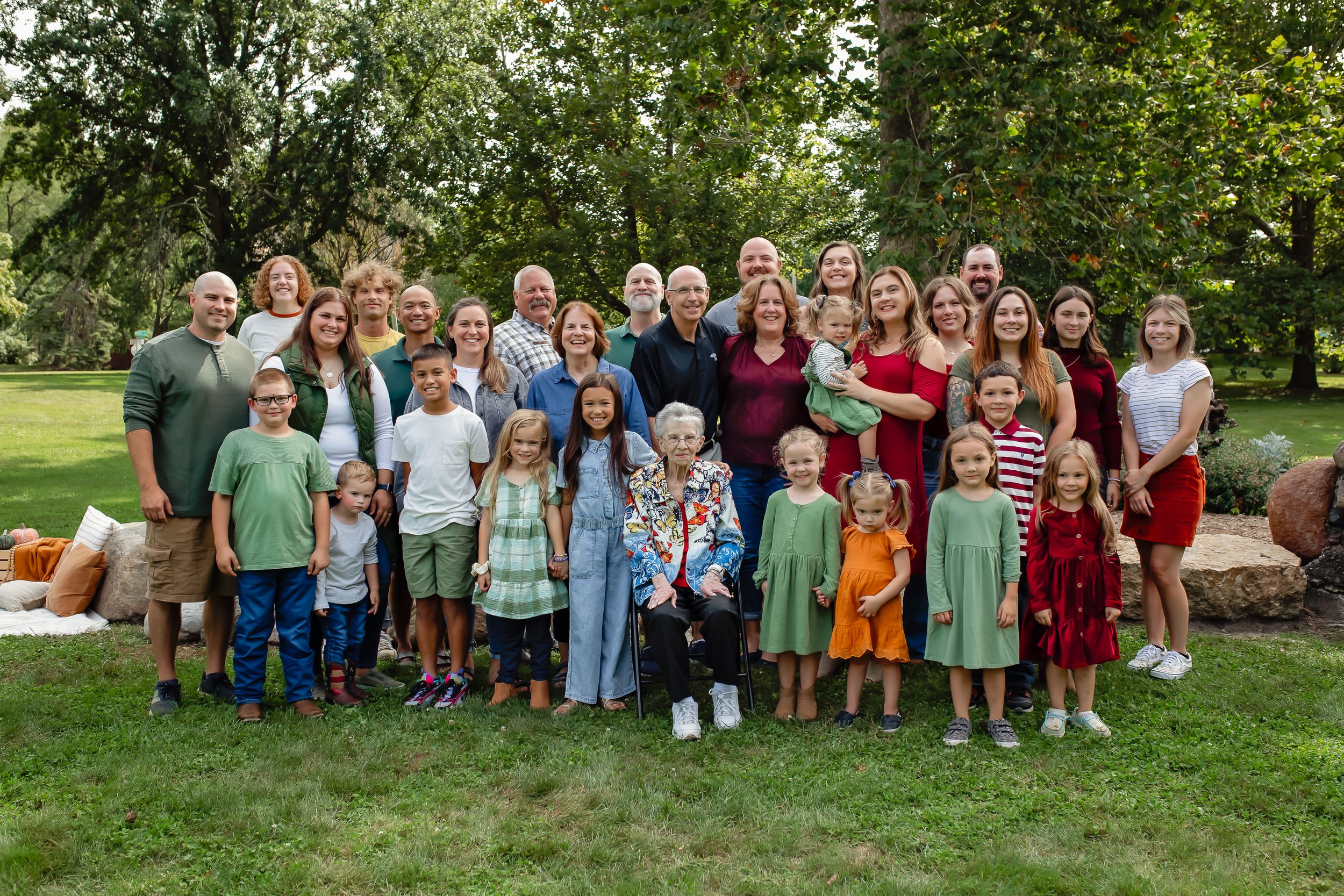 Large extended family group photo in a park with stone wall and greenery background.