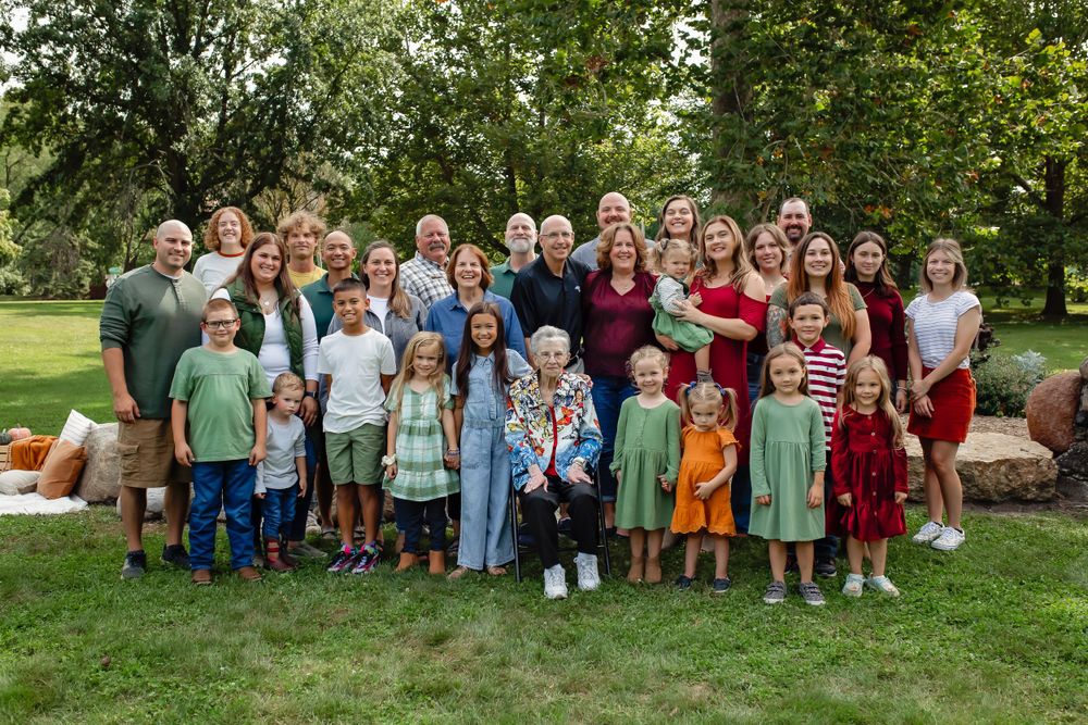 Large extended family group photo in a park with stone wall and greenery background.