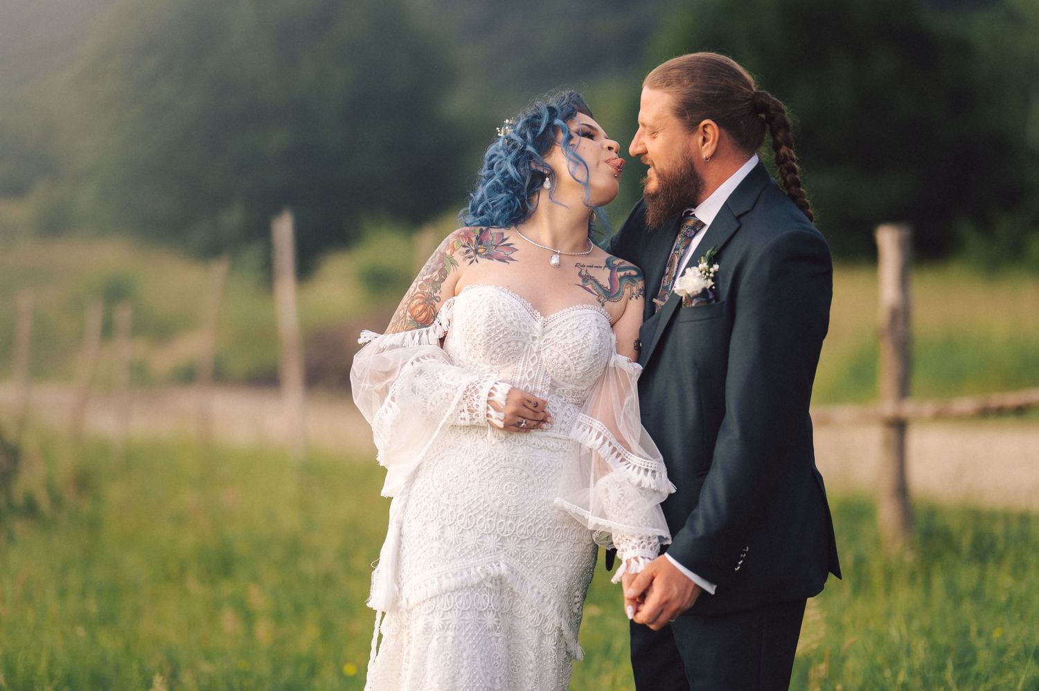 A romantic wedding photo of a couple standing in a lush green countryside meadow at sunset.