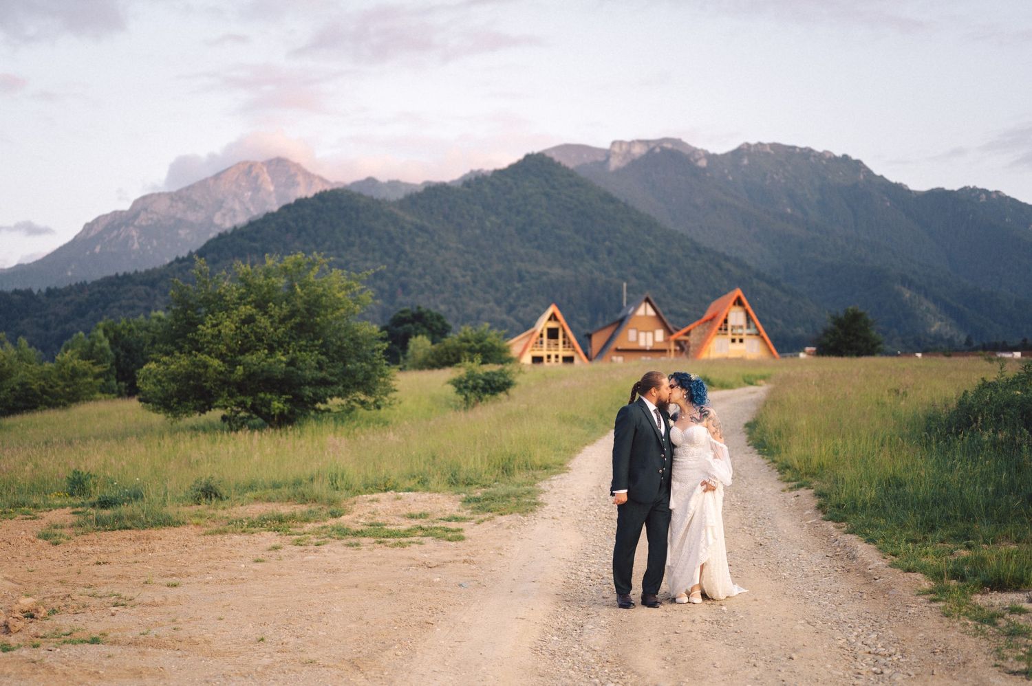 A couple stands on a dirt road with rustic A-frame cabins and majestic mountain peaks in the background.