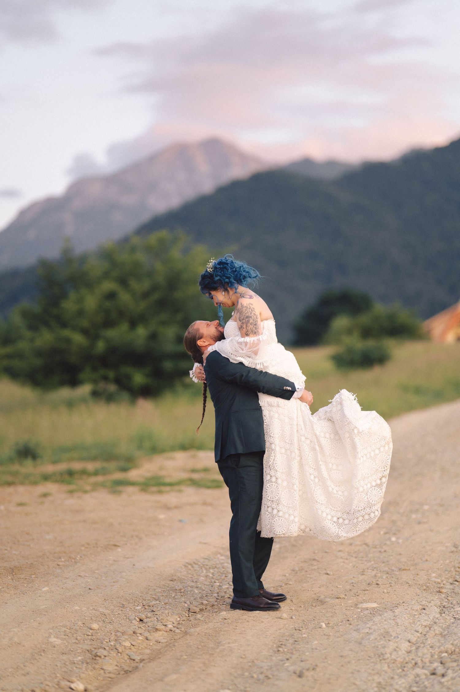 A couple shares a romantic embrace on a dirt road with majestic mountains and sunset sky in the background.