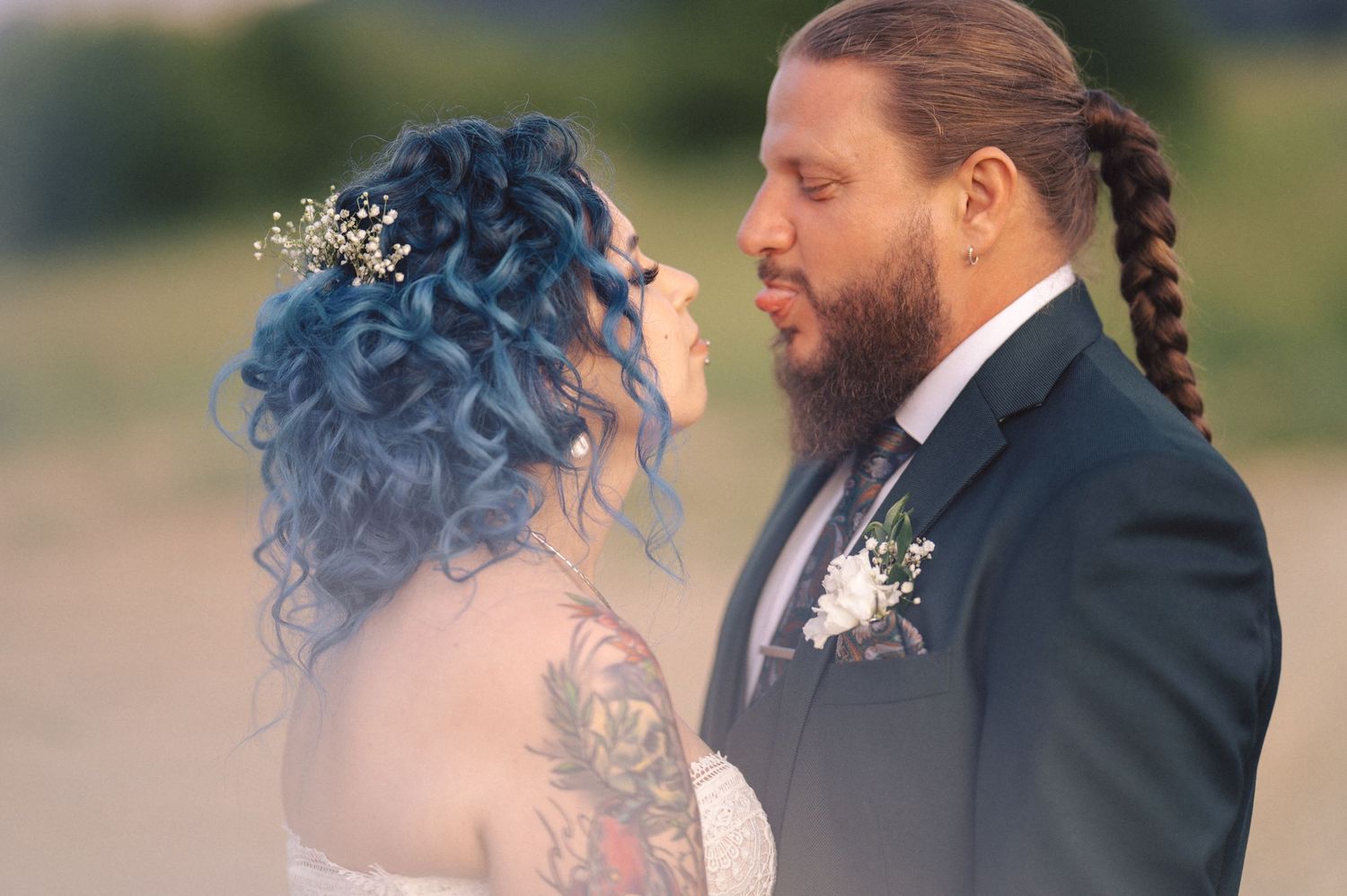 A couple in wedding attire share a romantic moment outdoors, with the bride wearing a unique blue hairstyle and white gown.