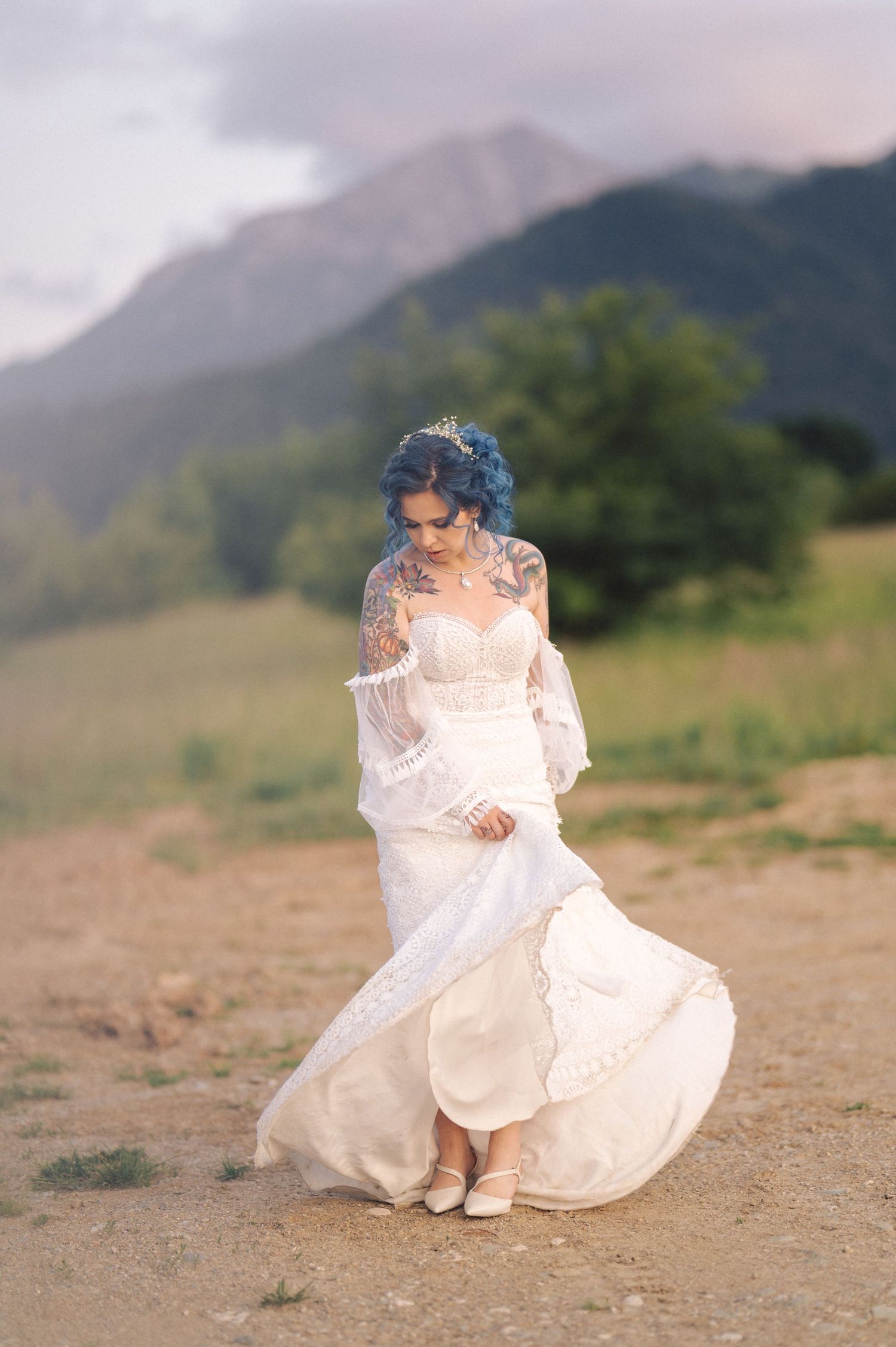 A bride in a flowing white dress sits gracefully on sandy ground with mountains visible in the misty background.