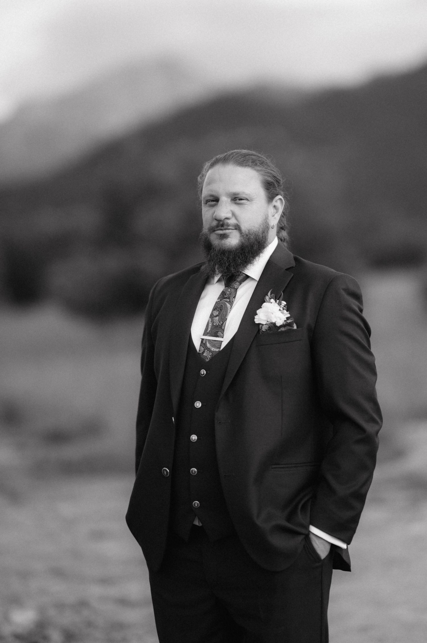 A bearded groom in a black three-piece suit and boutonniere stands outdoors against a mountain backdrop in black and white.