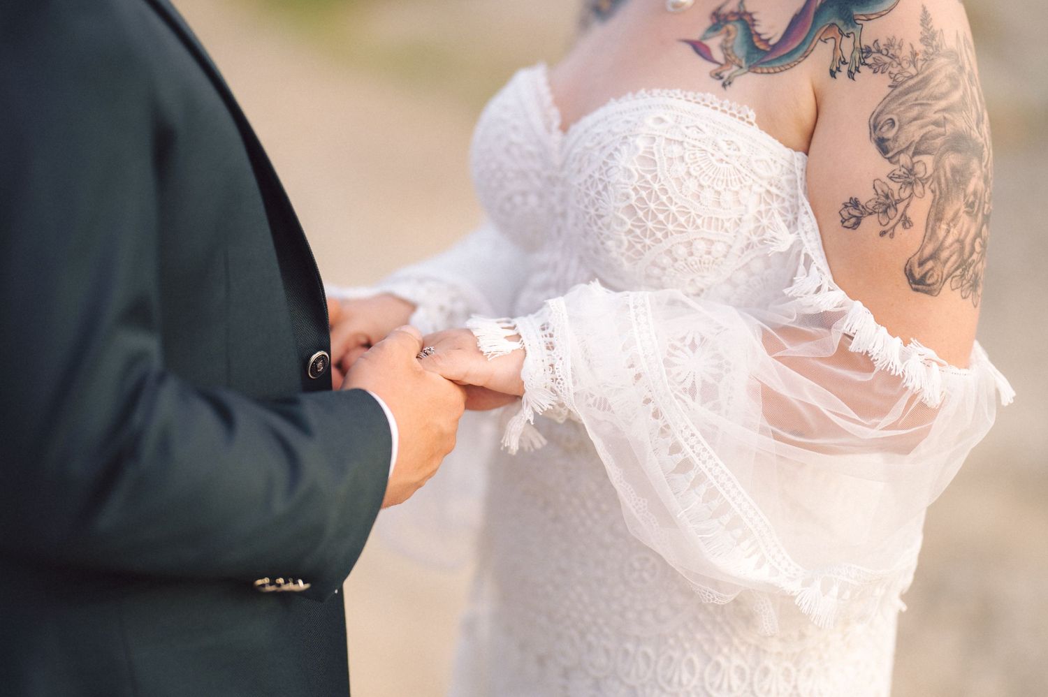 Close-up detail of a white lace wedding dress and veil with visible sleeve tattoo during outdoor ceremony.