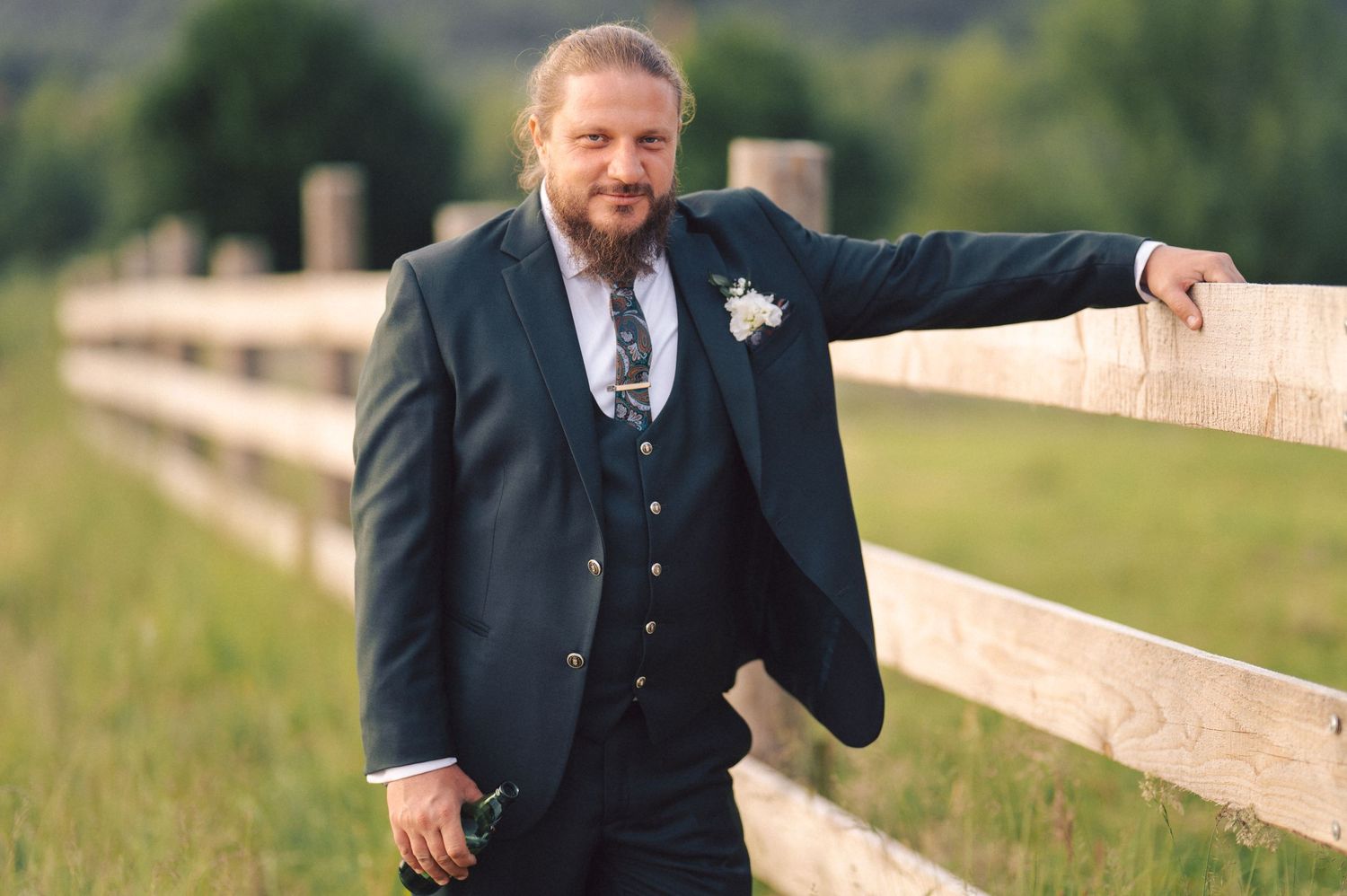 A groom in a navy suit stands in vibrant high grass by a wooden fence in a rural outdoor setting.