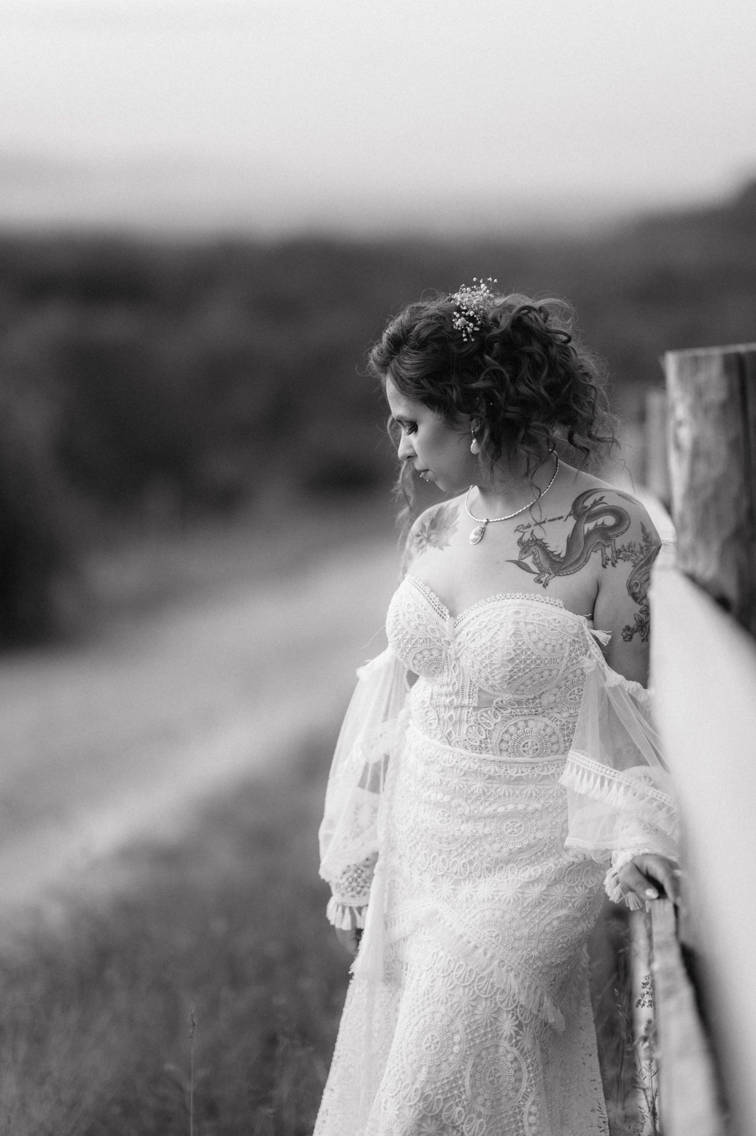 A bride in a delicate lace wedding dress with sheer sleeves and tattoos stands thoughtfully in a black and white portrait.