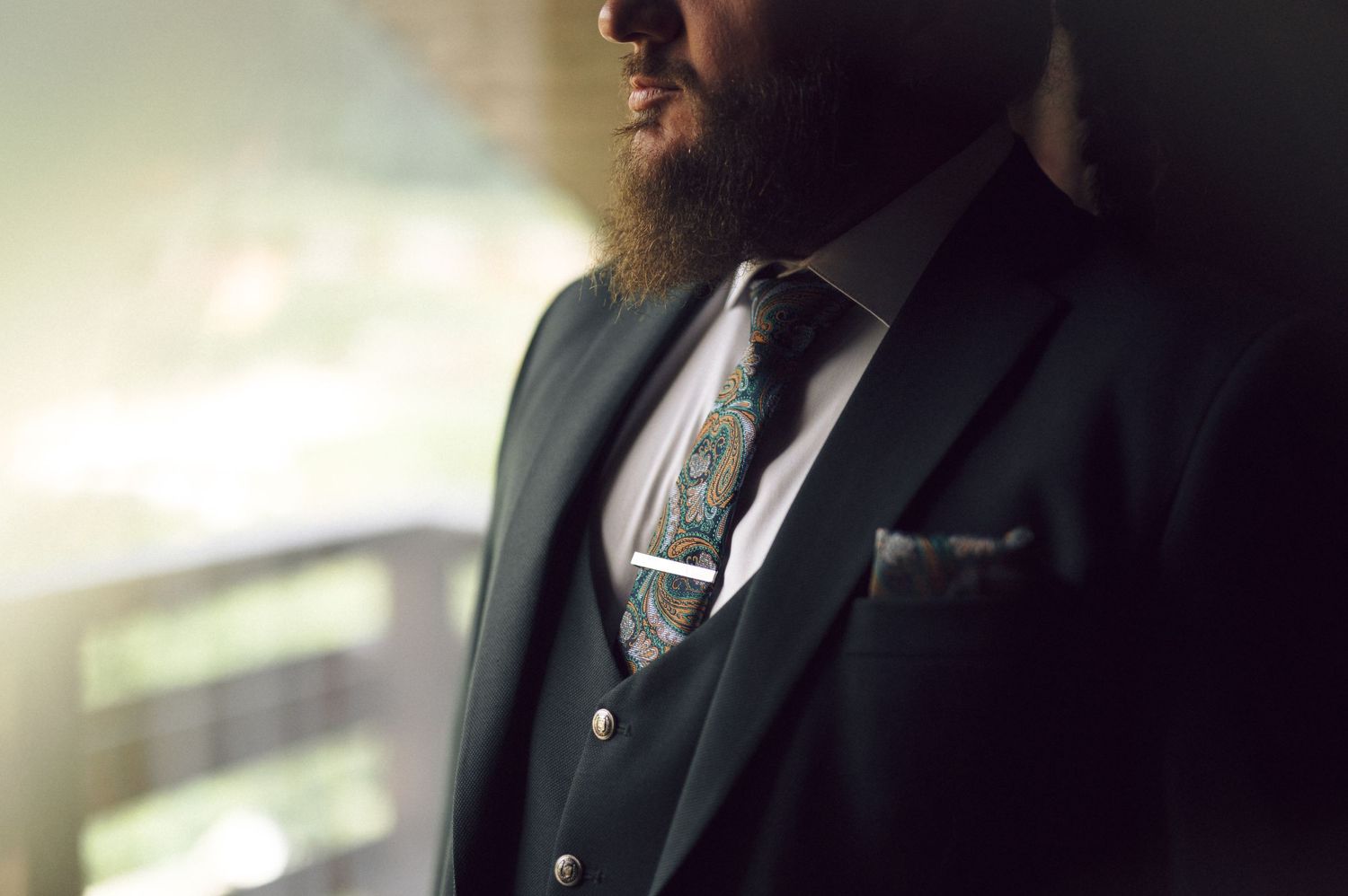 A close-up profile view shows a bearded individual wearing a black suit, vest and patterned tie in soft lighting.
