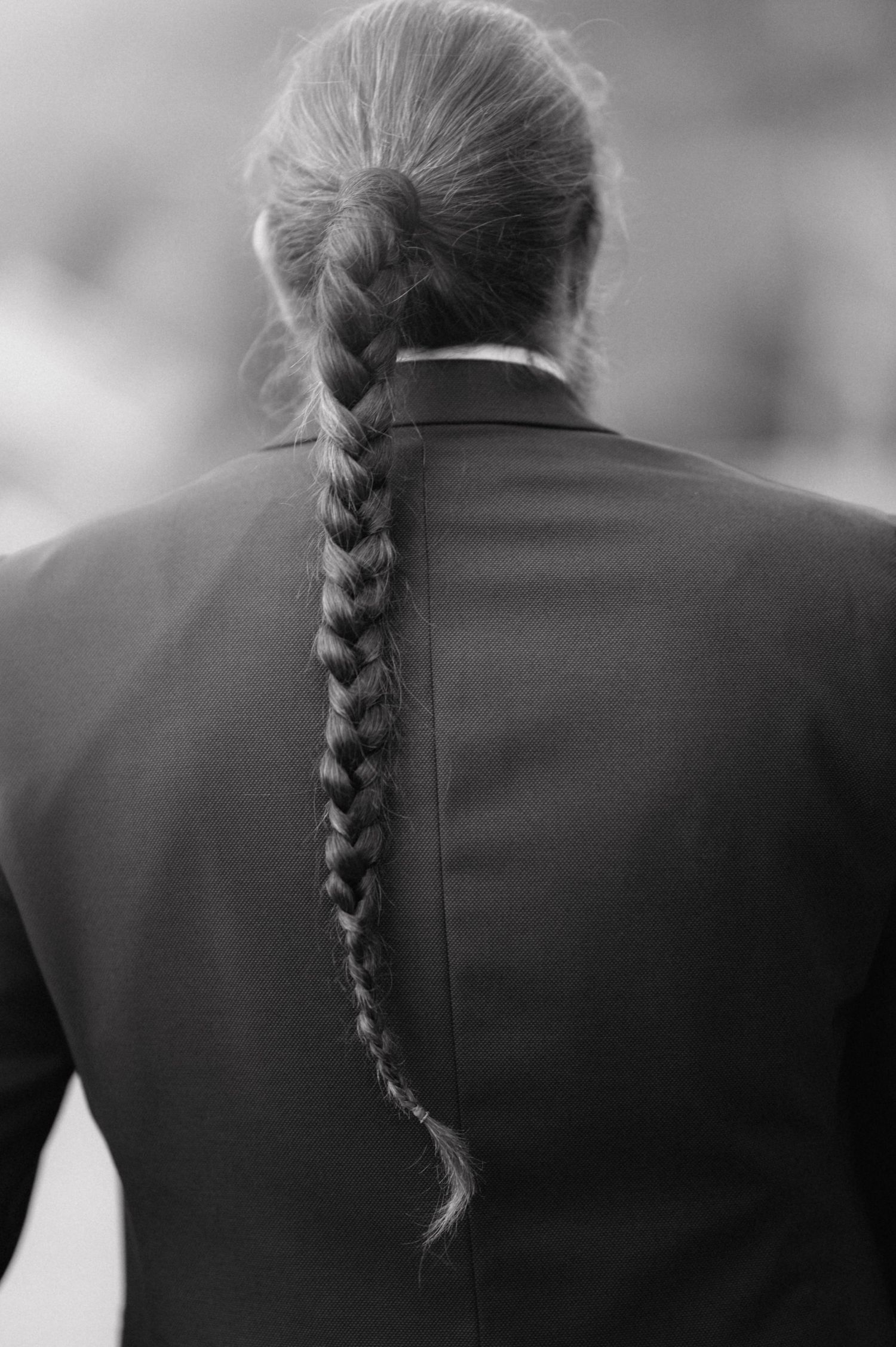 Black and white image shows the back of a person with a long single braid against a dark jacket and blurry background.
