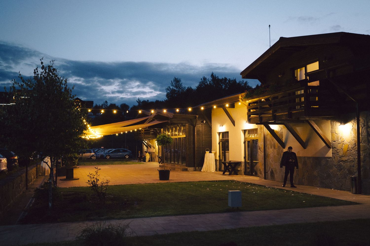 String lights illuminate a rustic building exterior with a warm glow during twilight as silhouettes gather on the patio.