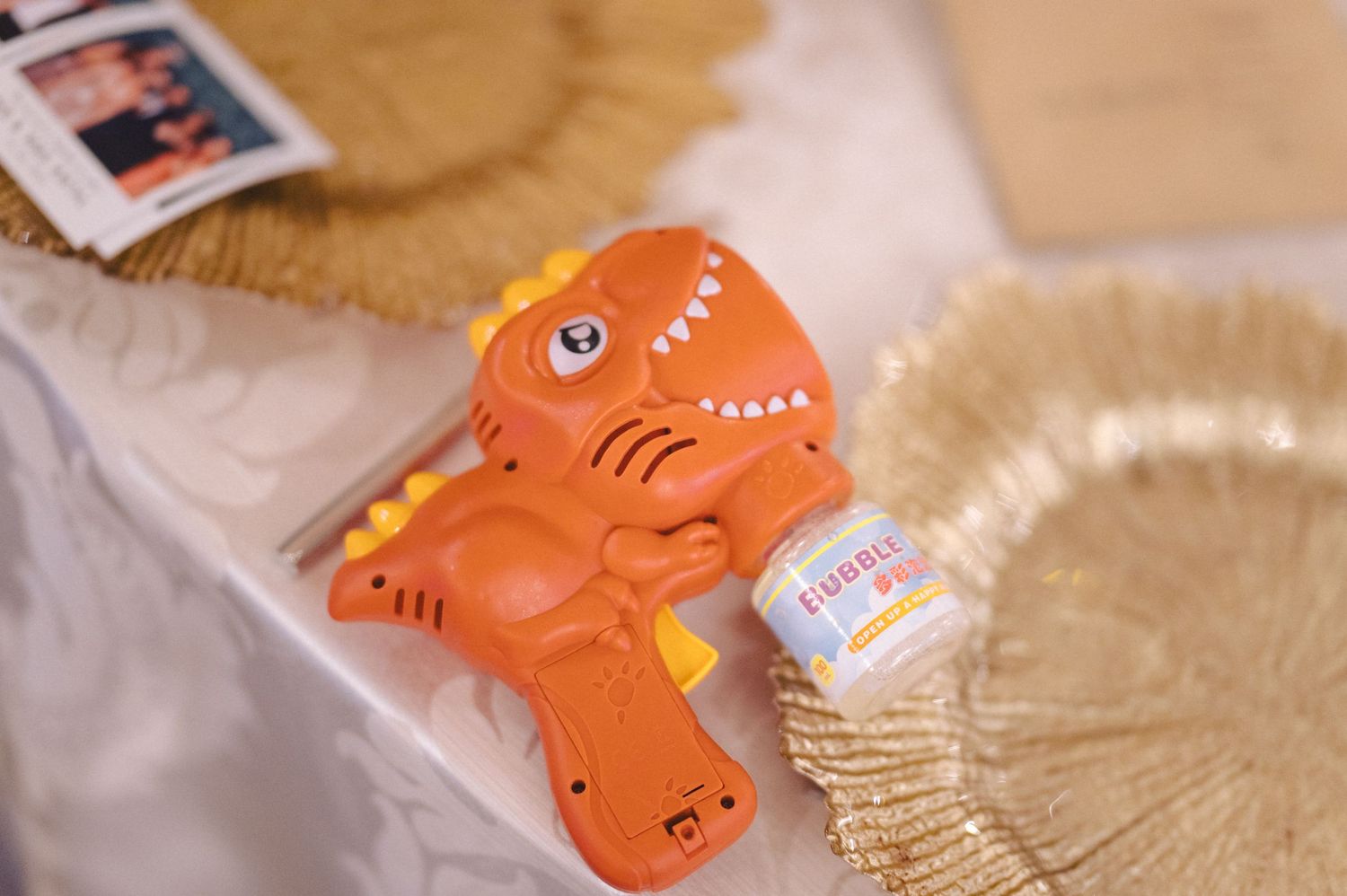 Orange dinosaur toy sits among gold sequins on a textured surface with metallic sparkle details.