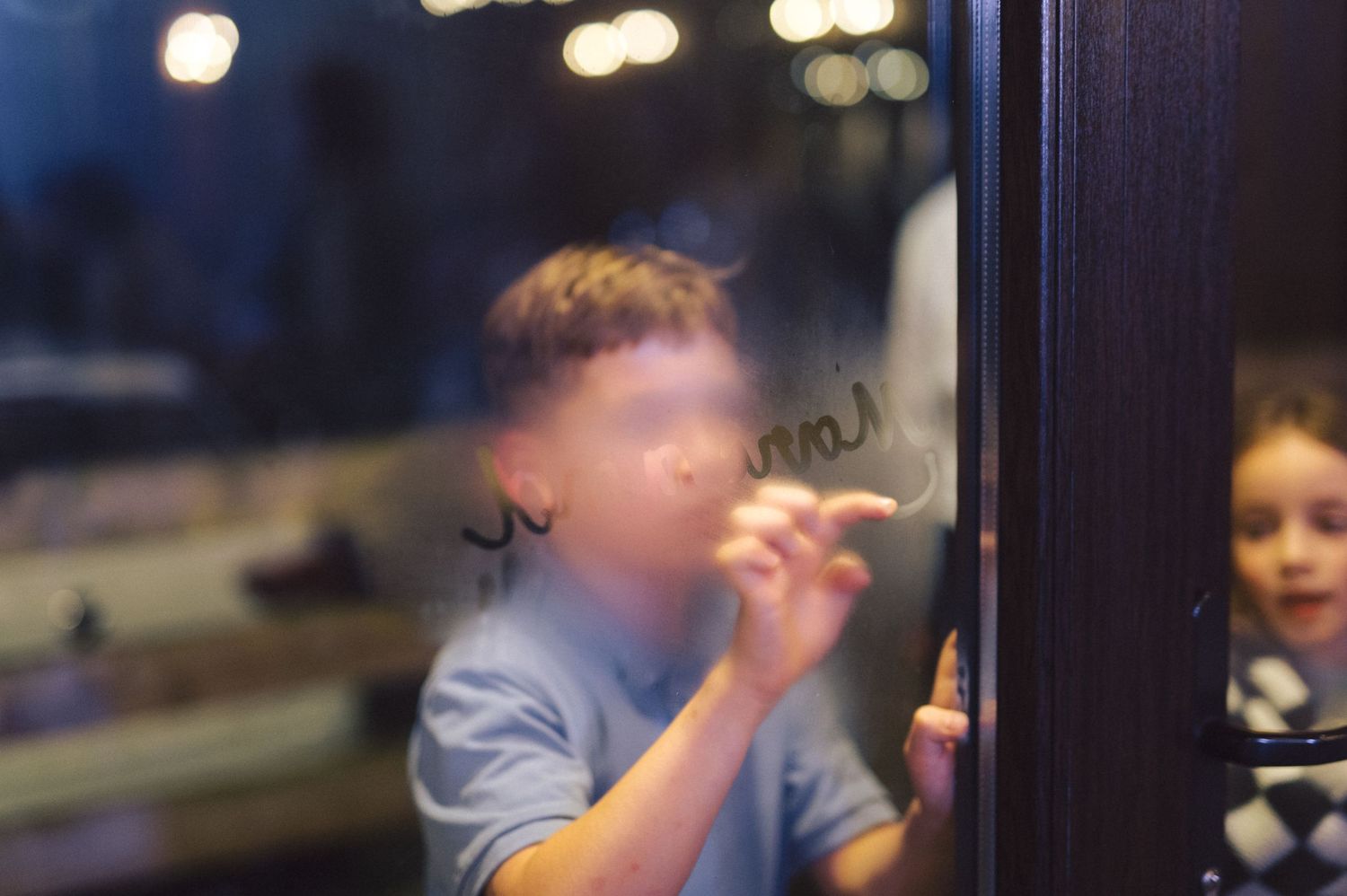 Someone with blonde hair smoking a cigarette while peering through a window with reflections and blurred city lights.
