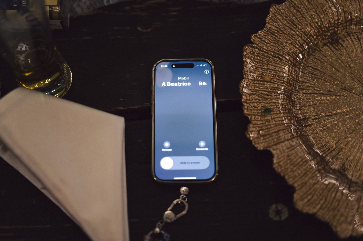 An iPhone displaying an app interface rests on a dark surface next to a light colored notebook and a golden textured item.