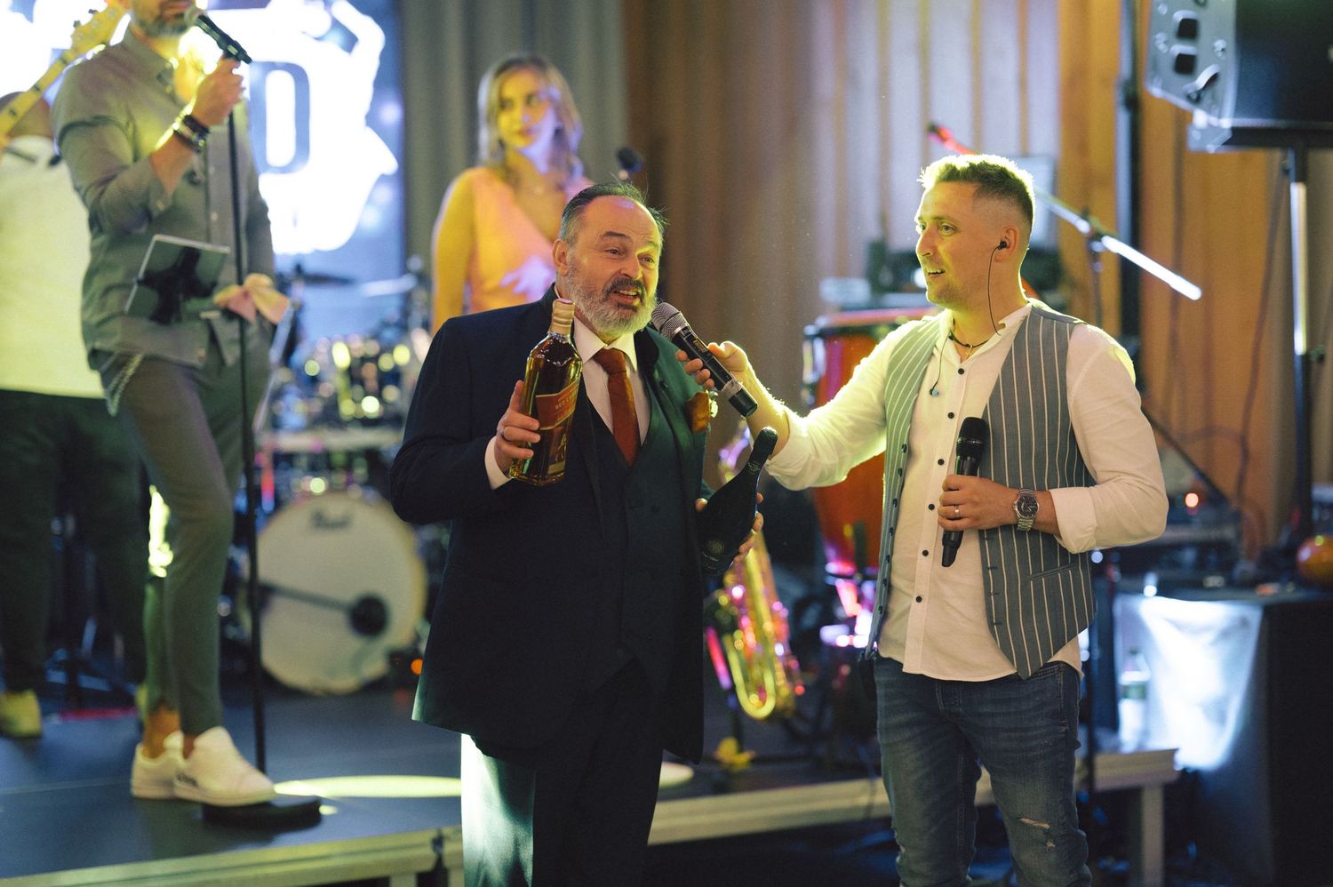 Musicians perform together on stage at an indoor venue with band instruments and lighting setup in the background.