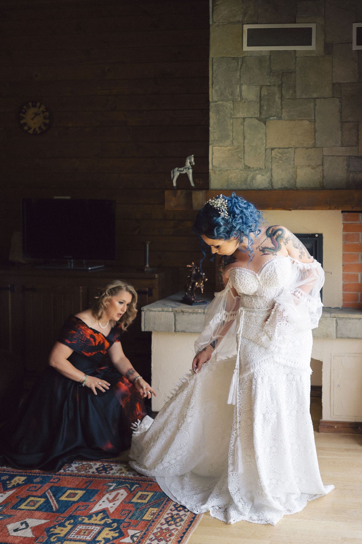 Two people help adjust an elegant white lace wedding dress in a room with stone walls and a decorative area rug.
