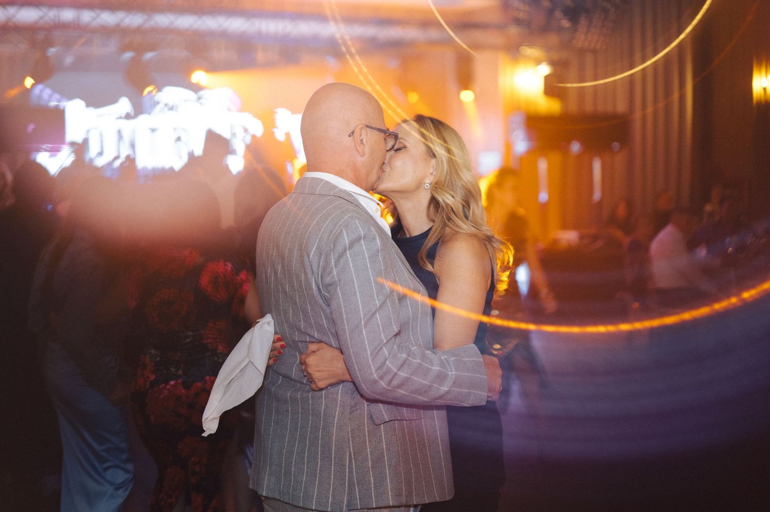 A couple embraces while dancing at an evening event with warm lighting and blurred motion effects in the background.