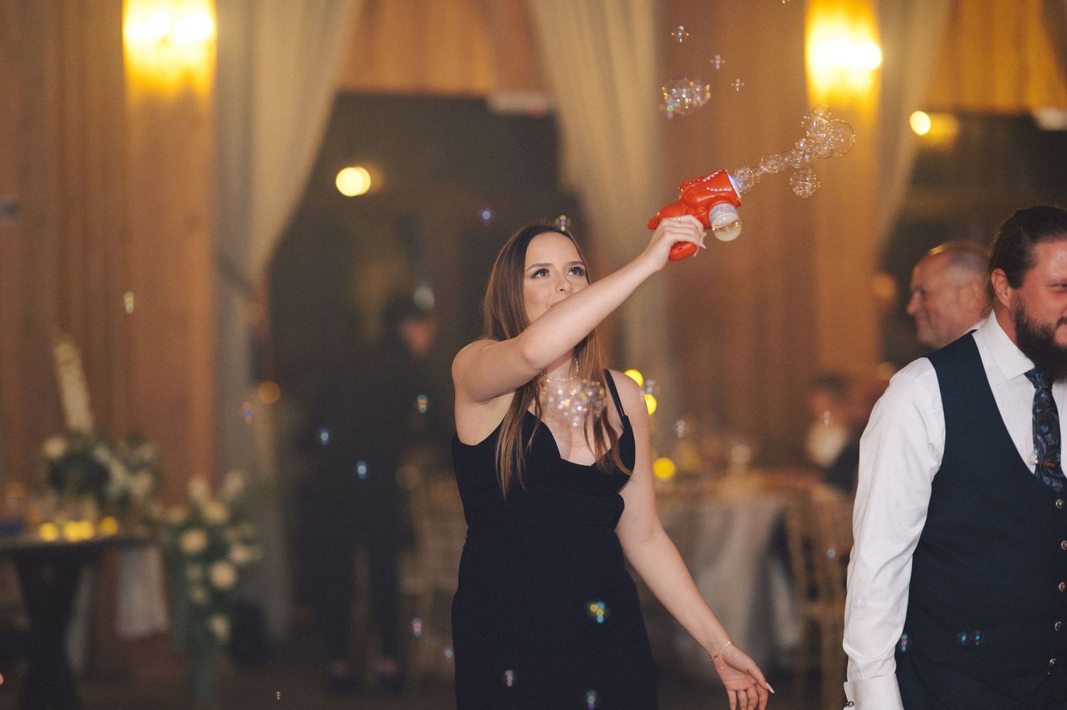 A party guest in a black dress sprays a champagne bottle at a elegant indoor reception venue during celebrations.
