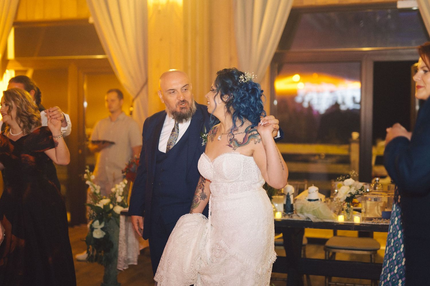 Couple dancing together at an elegant wedding reception with warm lighting and decorative table settings in the background.