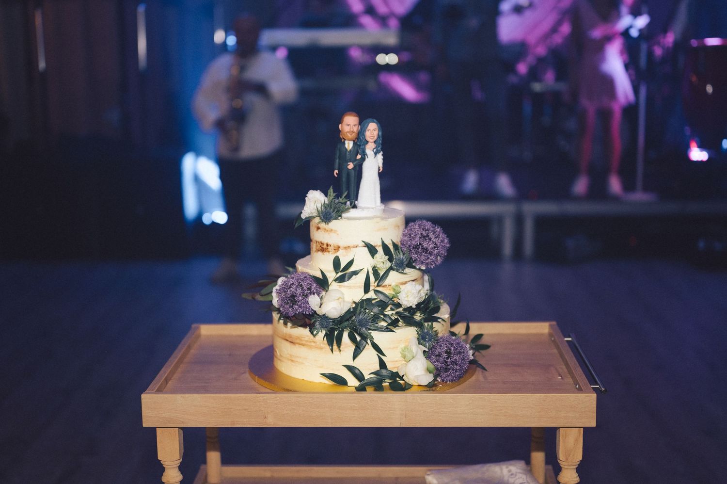 A three-tiered wedding cake with purple flowers and a traditional cake topper sits on a wooden table in dim lighting.