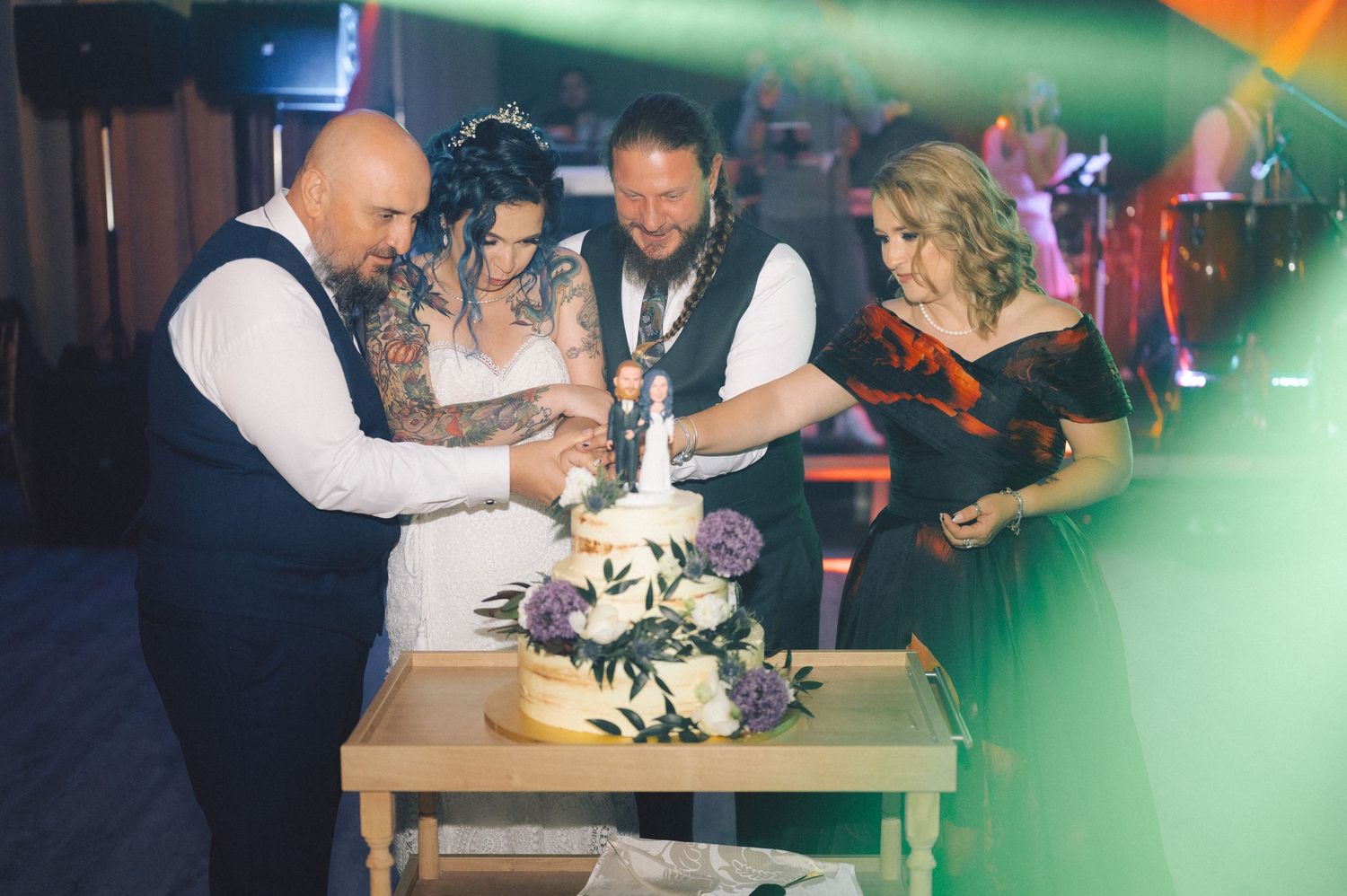 A group gathers around a wedding cake at a reception venue with colorful lighting and festive atmosphere.