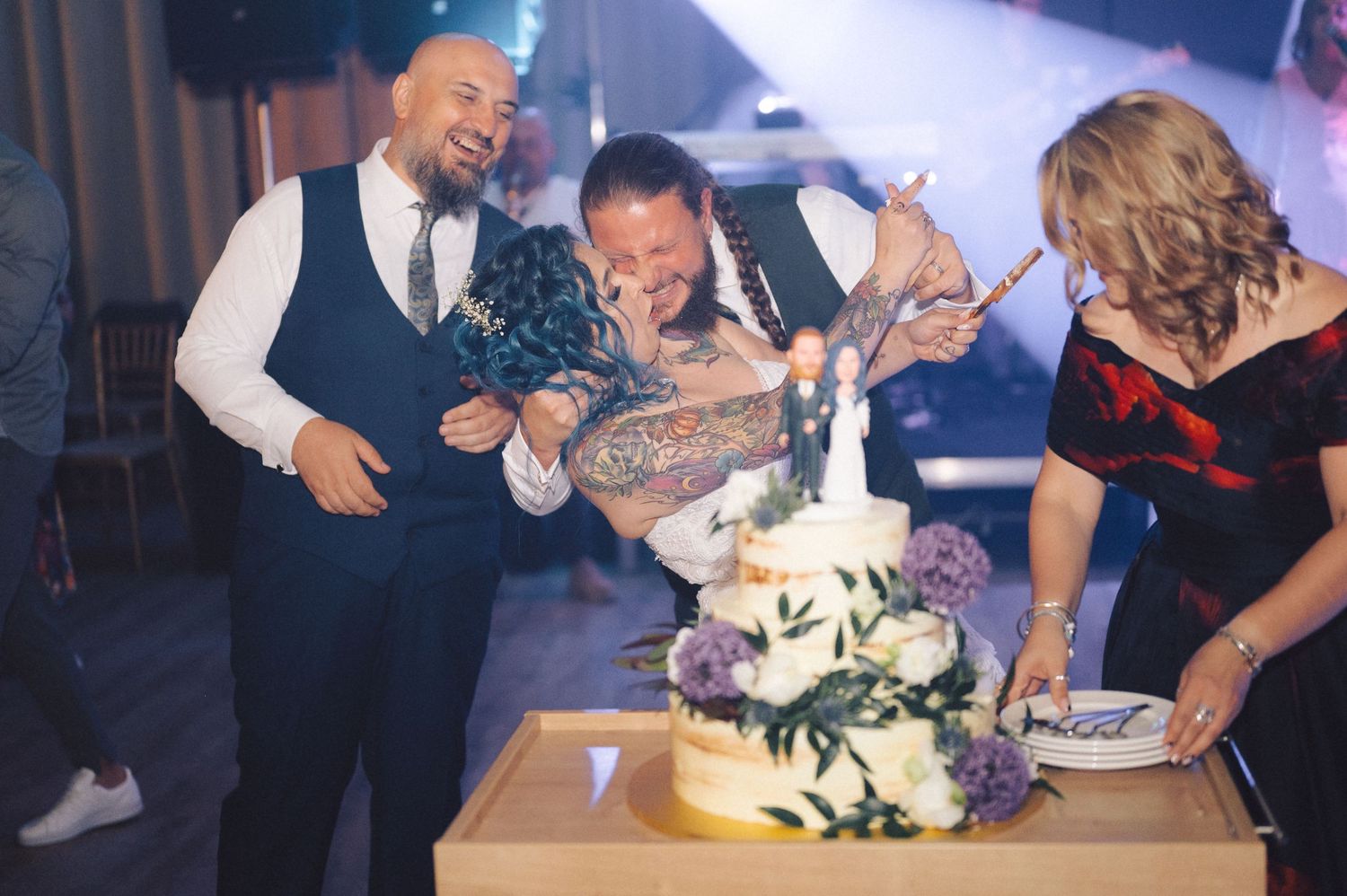 Wedding party celebrates during cake cutting ceremony as guests laugh and party at an indoor reception.